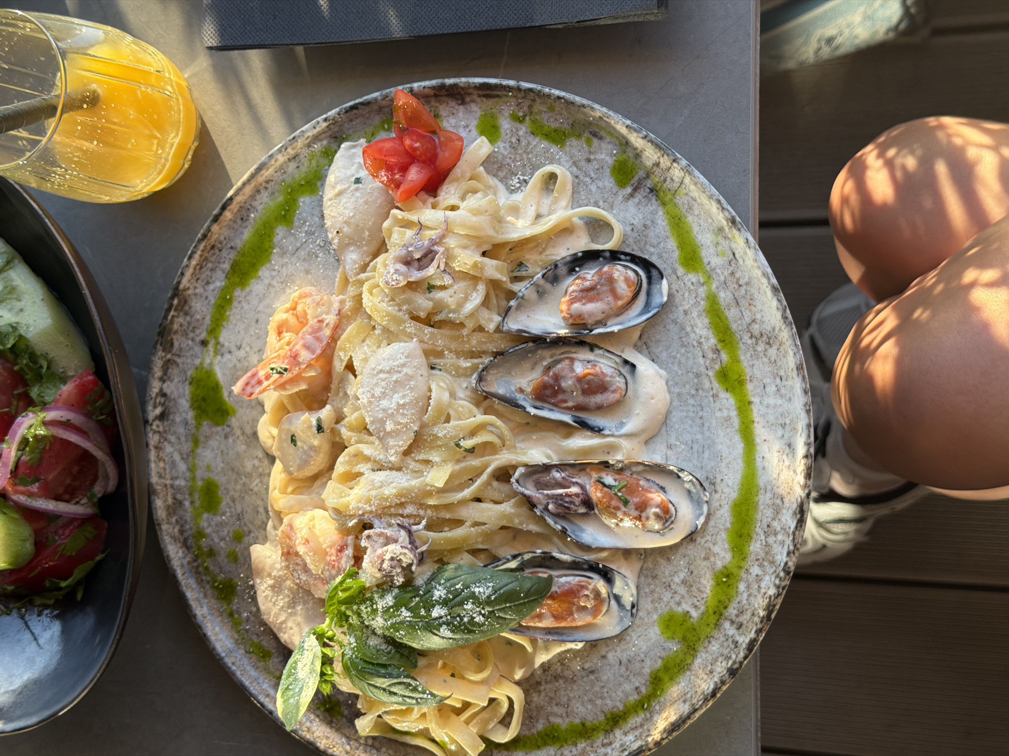 The image depicts a plate of seafood pasta, including mussels, shrimp, squid, and tomatoes, garnished with basil and served on a textured plate with green sauce. The pasta is creamy. A glass of orange juice is beside the plate. A side dish of salad is also visible. A child is sitting in a high chair beside the plate. The location is in Rafailovići, Montenegro. The environment suggests an outdoor dining setting.