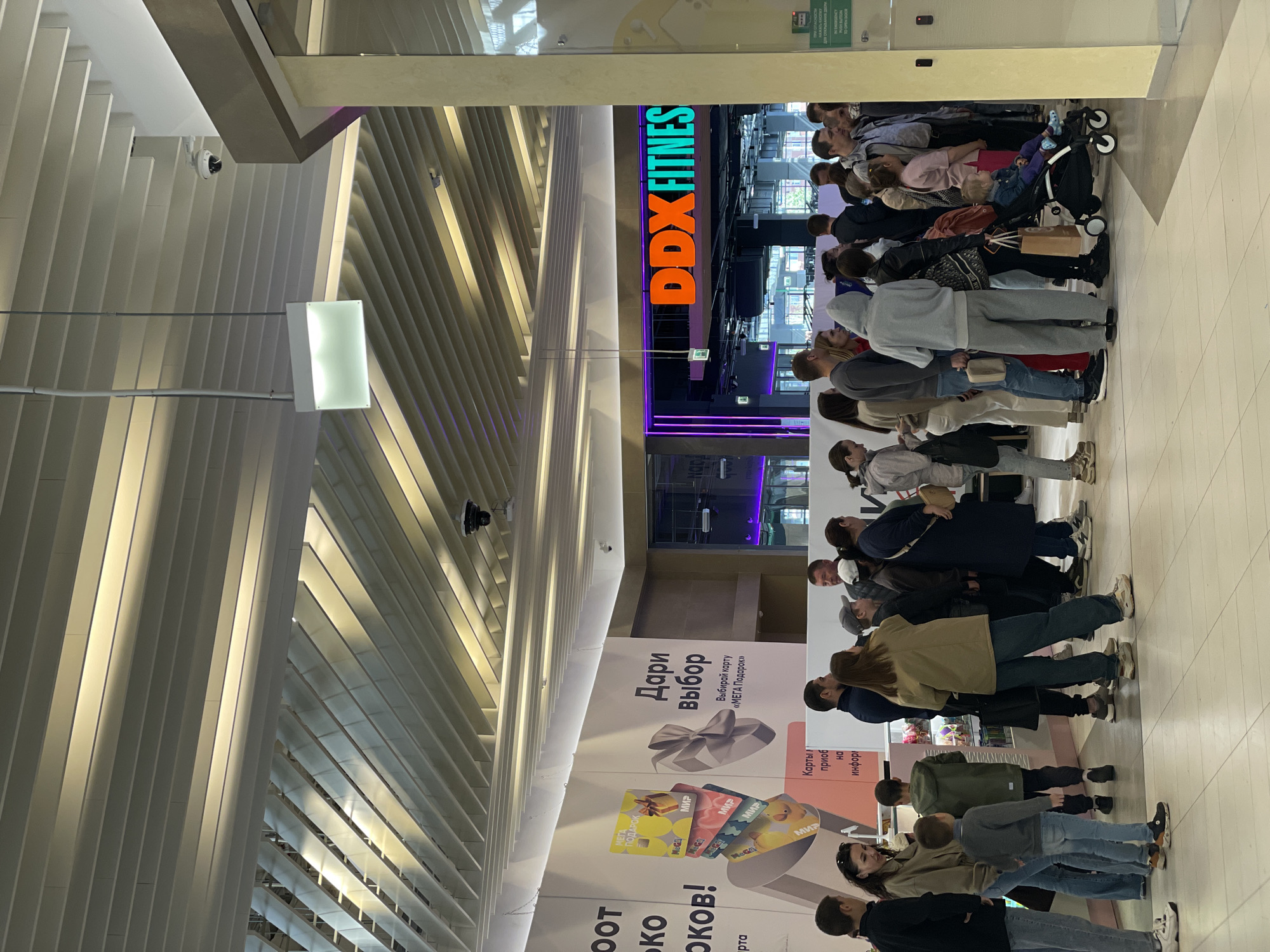 The image shows a long line of people waiting outside a DDX Fitness center. The scene is located in a shopping mall in Kazan, Russia. The people in line vary in age and are dressed in casual clothing. A sign advertises gift cards from "MEGA." The lighting is bright, suggesting an indoor location. A stroller is present in the line.
