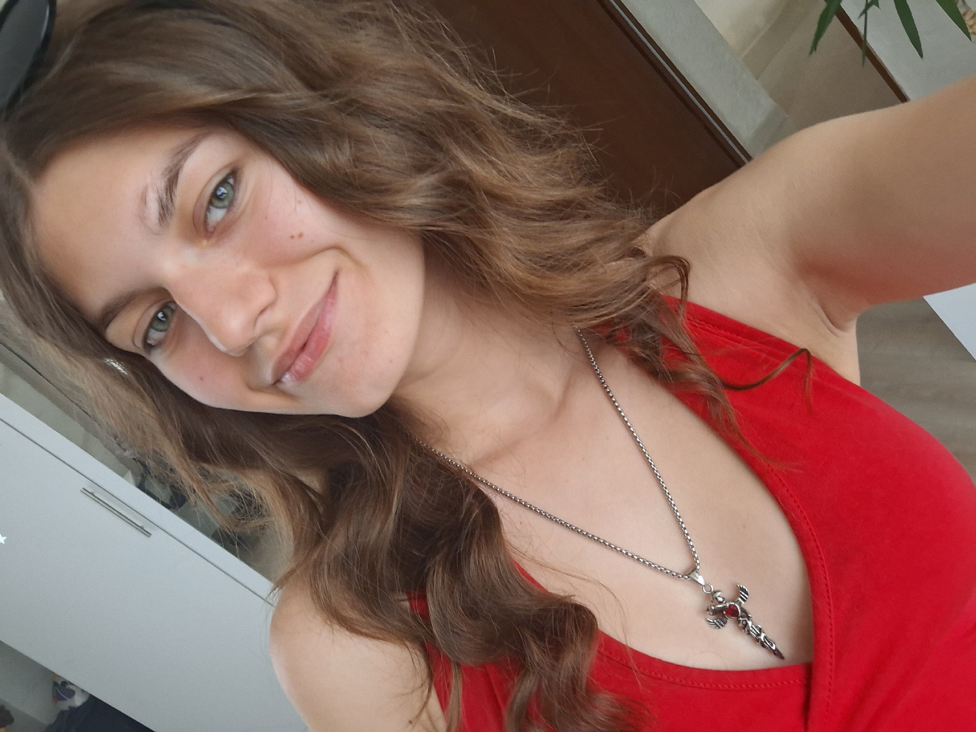 The image is a close-up, selfie-style shot of a young woman with wavy brown hair and green eyes. She is wearing a red tank top and a silver chain necklace with a cross-shaped pendant featuring a red gem. She is smiling and looking at the camera. The background appears to be indoors, with a light-colored cabinet or wardrobe visible on the left and a wooden doorframe on the right. A blurry green plant is also visible in the upper right corner. The image does not contain any information about the location in the city of Panagyurishte, Bulgaria.