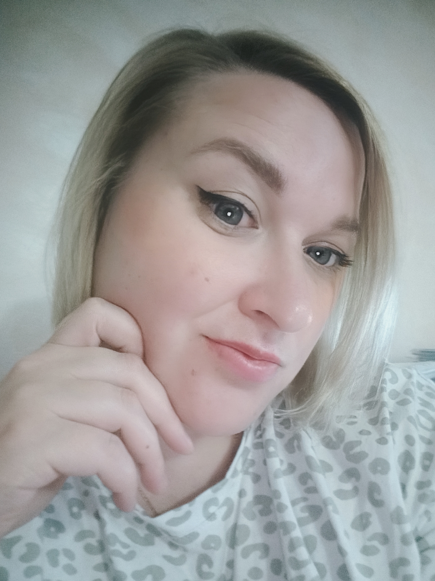 A close-up shot shows a fair-skinned woman with light blonde hair and gray eyes. She has a light makeup application with a defined eyeliner. Her right hand is raised, with her fingers gently touching the side of her face. She is wearing a light-colored top with a gray leopard print pattern. The background is a plain light-colored wall. The location is unknown.