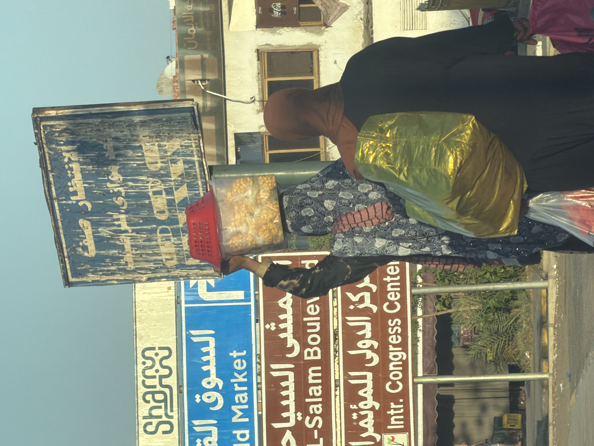 In Sharm El Sheikh, Egypt, two individuals are visible on a street. One person, wearing a dark abaya and a brown hijab, carries a large shiny gold bag over their shoulder and a pink bag in their hand. The other person, wearing a patterned abaya and hijab, is partially obscured and carries a red basket on their head filled with yellow popcorn kernels.
The background features several signs. One sign, partially obscured and weathered, has blue and yellow text. Another prominent sign is blue with white Arabic text and the words "Old Market" in English. Below that, a brown sign reads "L-Salam Boulev" in white English text, and another brown sign indicates "Intr. Congress Center" in English. Buildings with windows are visible behind the signs. The scene appears to be a street in a commercial area, possibly near a market or event center.