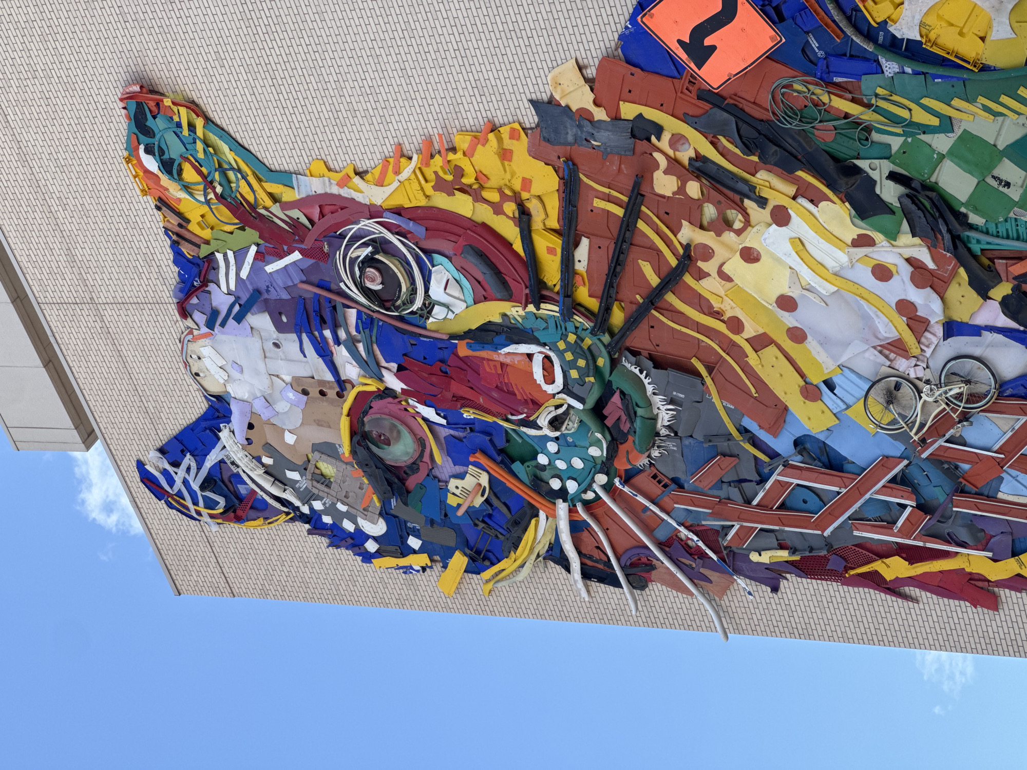 This is a close-up, low-angle outdoor photograph of a large, colorful mural depicting a cat's head. The mural is made from recycled materials and is mounted on the side of a light-colored brick building. The cat's head is a mosaic of various discarded items, including plastic pieces, metal fragments, and what appear to be parts of signs or traffic devices. The cat's eyes are particularly striking, constructed with layered circular elements and wires. The mural is vibrant with a wide range of colors, including blues, yellows, reds, greens, and purples. A clear blue sky with a few wispy white clouds is visible in the background to the left of the building. Parts of the mural include a small white bicycle and an orange traffic sign with a black arrow. This mural is located in El Paso, Texas, United States.