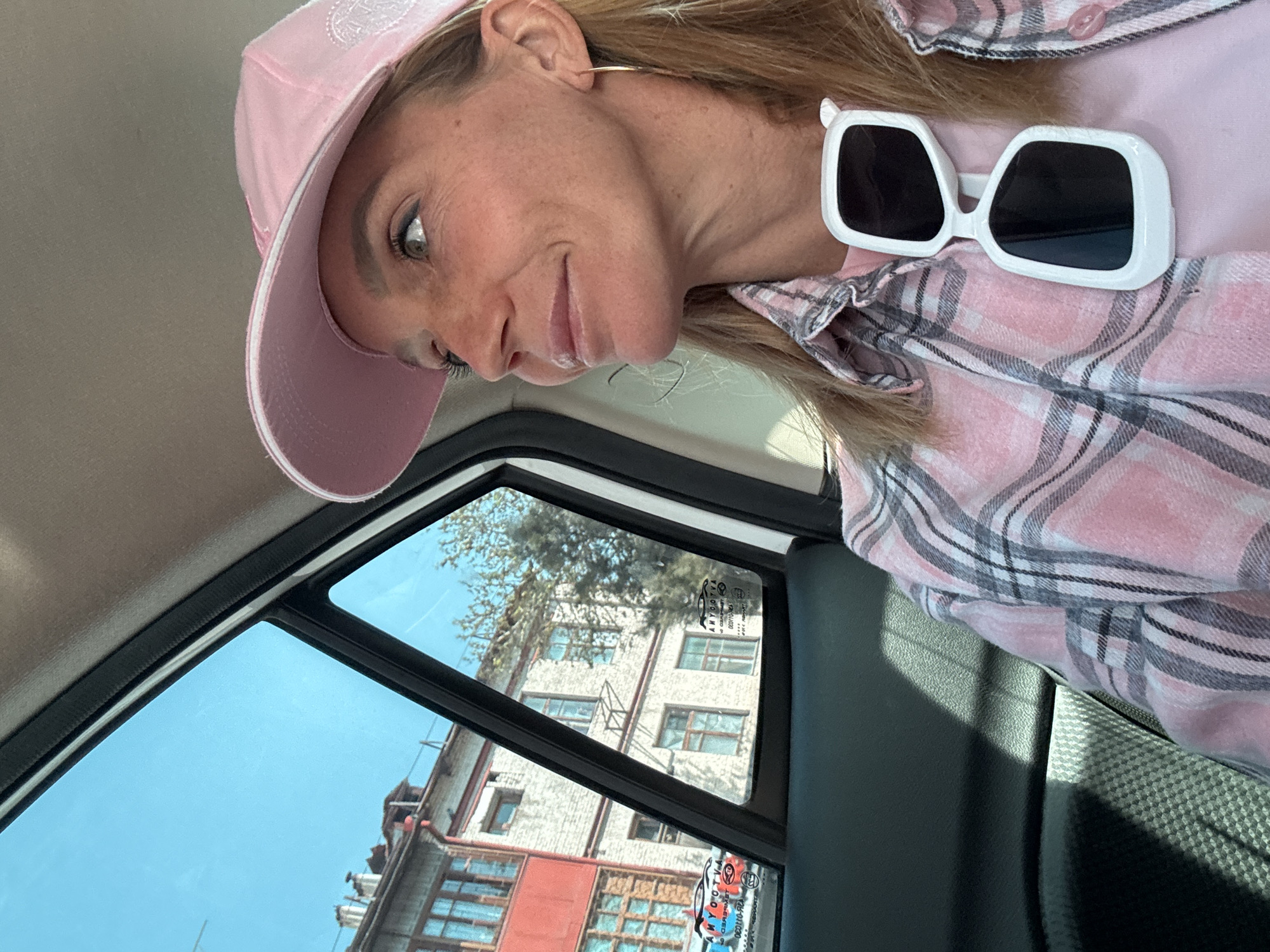 A medium shot shows a woman wearing a pink baseball cap and pink plaid shirt in what appears to be a vehicle. She is looking down and slightly towards the camera. A pair of white, square sunglasses are nestled in her shirt near her shoulder. The window of the vehicle provides a view of a multi-story building across the street in Tashkent, Uzbekistan.
