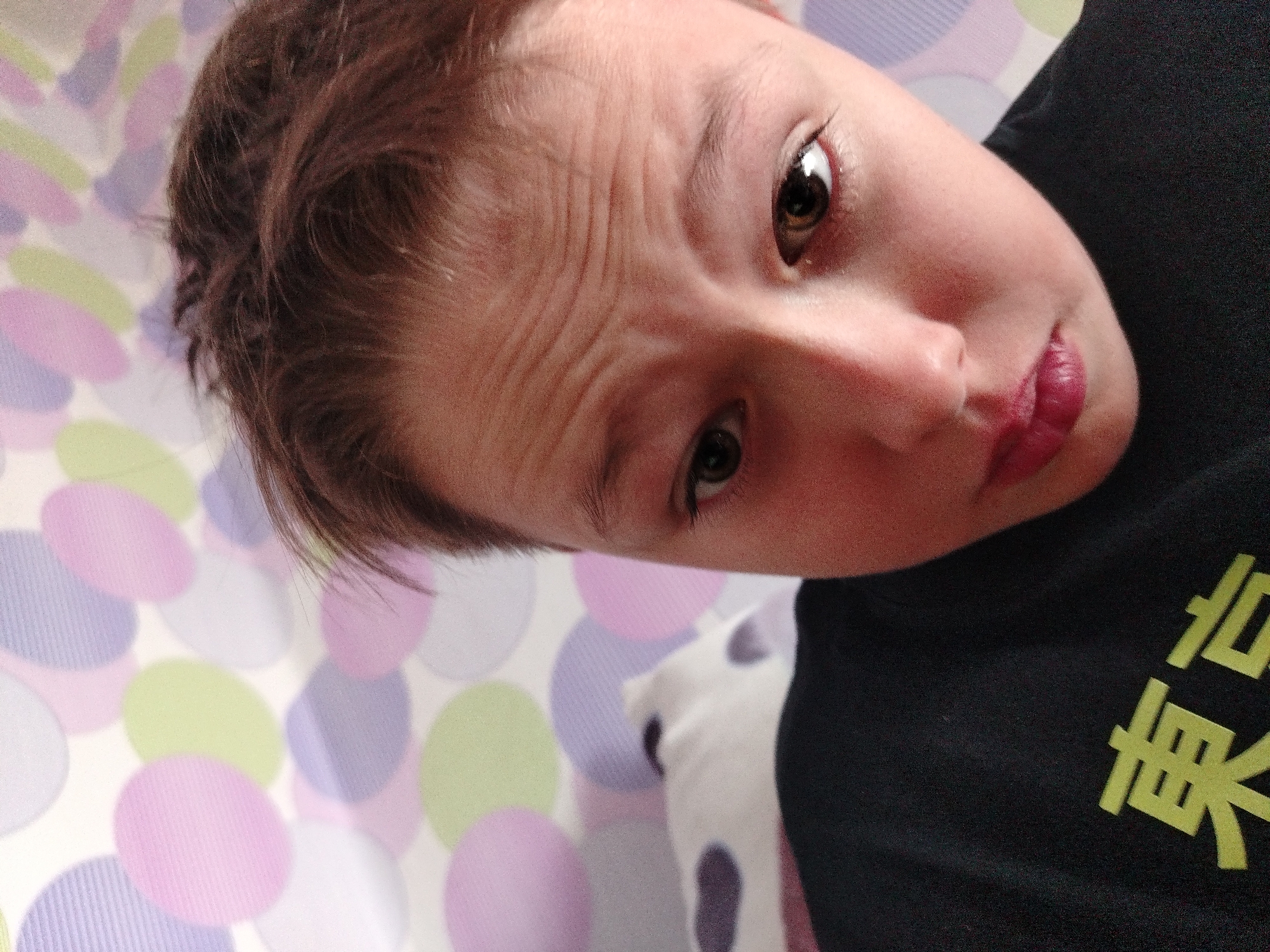 The image shows a close-up, angled shot of a child's face, mostly obscuring the background which appears to be a wall with a polka dot pattern in pastel colors. The child's head is tilted, and their brown eyes are looking towards the camera. Their forehead shows distinct wrinkles. The child is wearing a black t-shirt with yellow characters on it, which appear to be in an East Asian script. The lighting is dim, casting shadows on the child's face. The image is a selfie taken indoors, and does not contain information about the city of Chaikovsky, Russia.