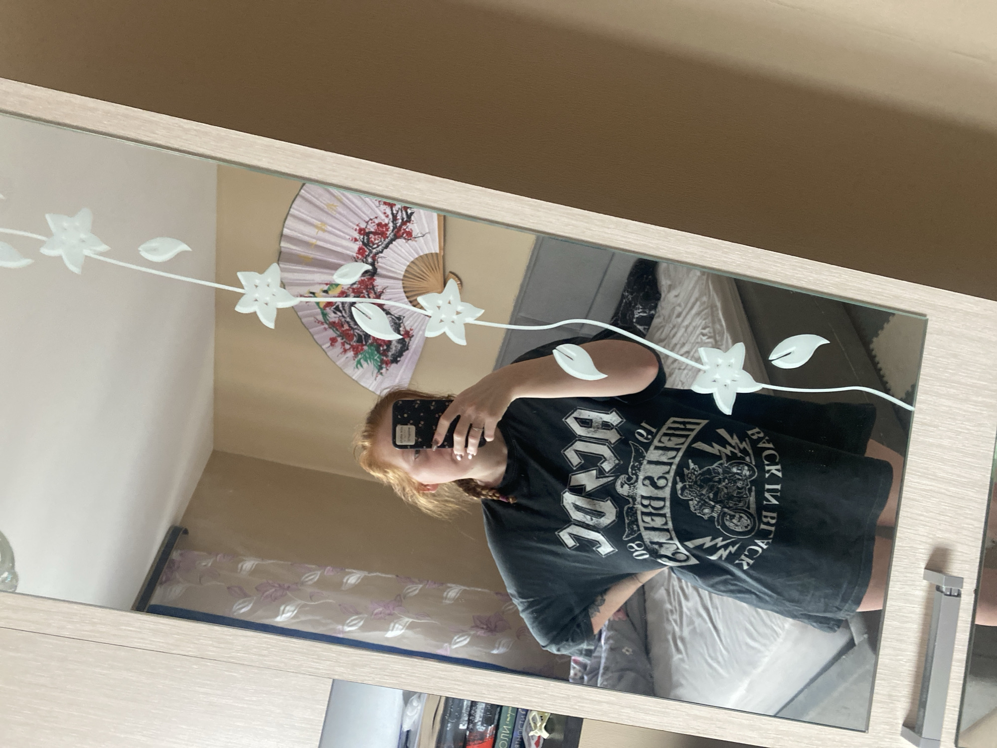 The image depicts a reflection in a mirror showing a person with red hair and braids wearing a black AC/DC t-shirt, holding a black phone with a patterned case to take a selfie. Behind the person in the reflection is a bed covered with a white blanket and grey pillows. On the wall is a decorative fan with a floral pattern. Below the mirror is a shelf with books. The location is unclear from the image and cannot be determined to be Yekaterinburg, Russia.