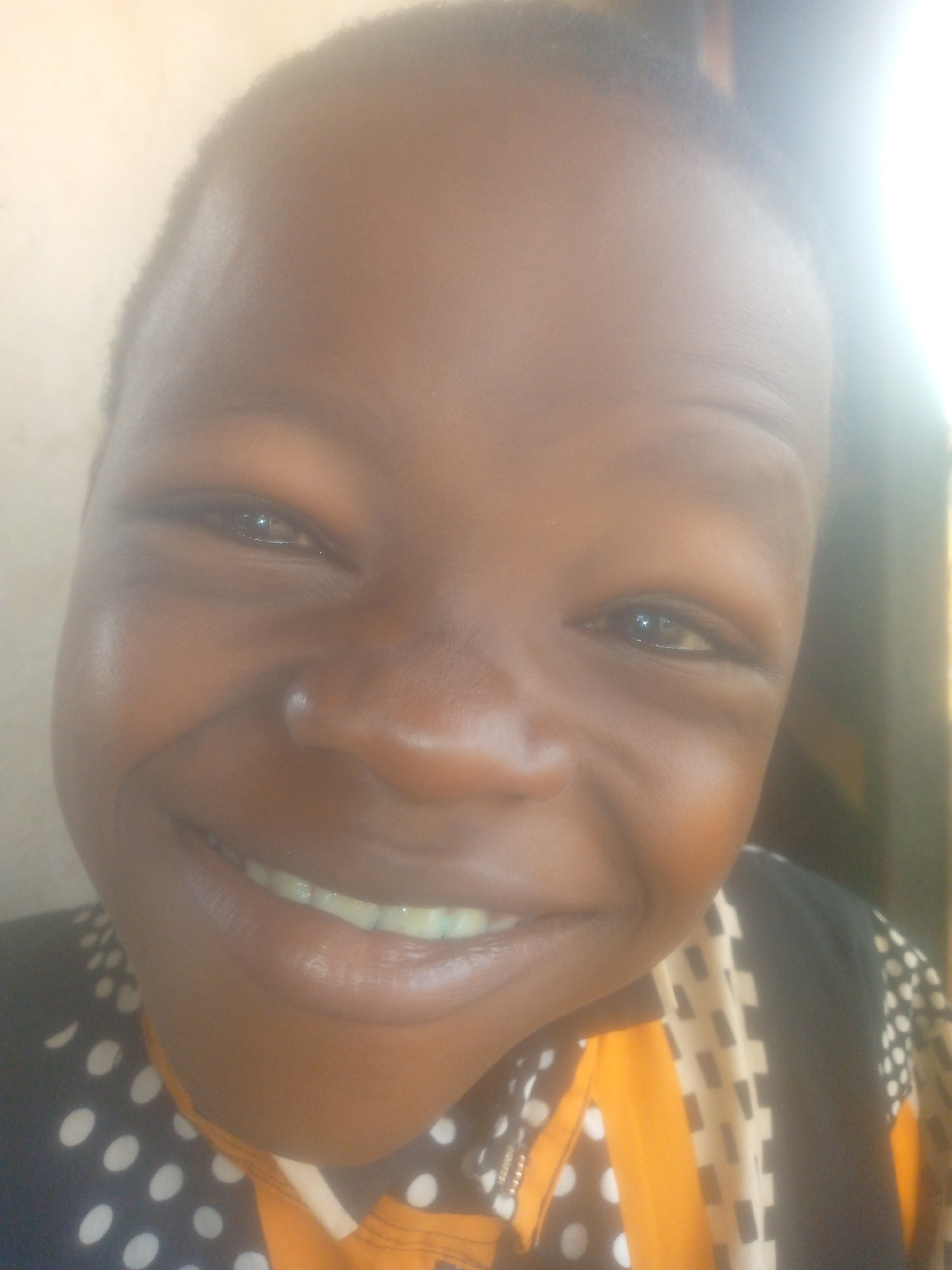 A close-up shot shows the face of a young person with dark skin smiling broadly, revealing their teeth. They wear a shirt with a mixed pattern of polka dots and a geometric design with orange and black elements. The background is blurred, suggesting an indoor environment with a wall or surface behind the person. The location is Unknown, Unknown.