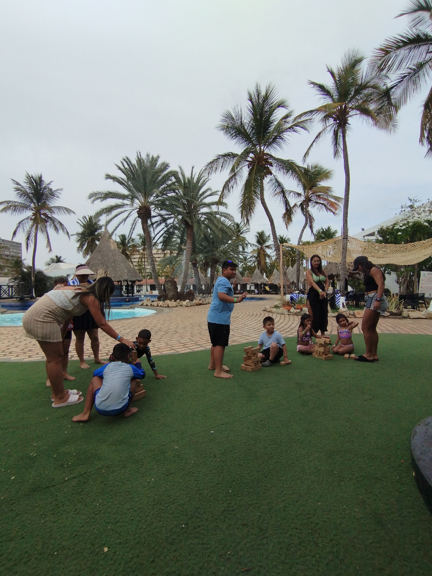 The image depicts a group of children and adults playing Jenga on a green lawn area, likely artificial turf, outdoors in Porlamar, Venezuela. In the background are palm trees, thatched-roof structures, a swimming pool, and buildings. The adults are interacting with the children, guiding or observing their game.