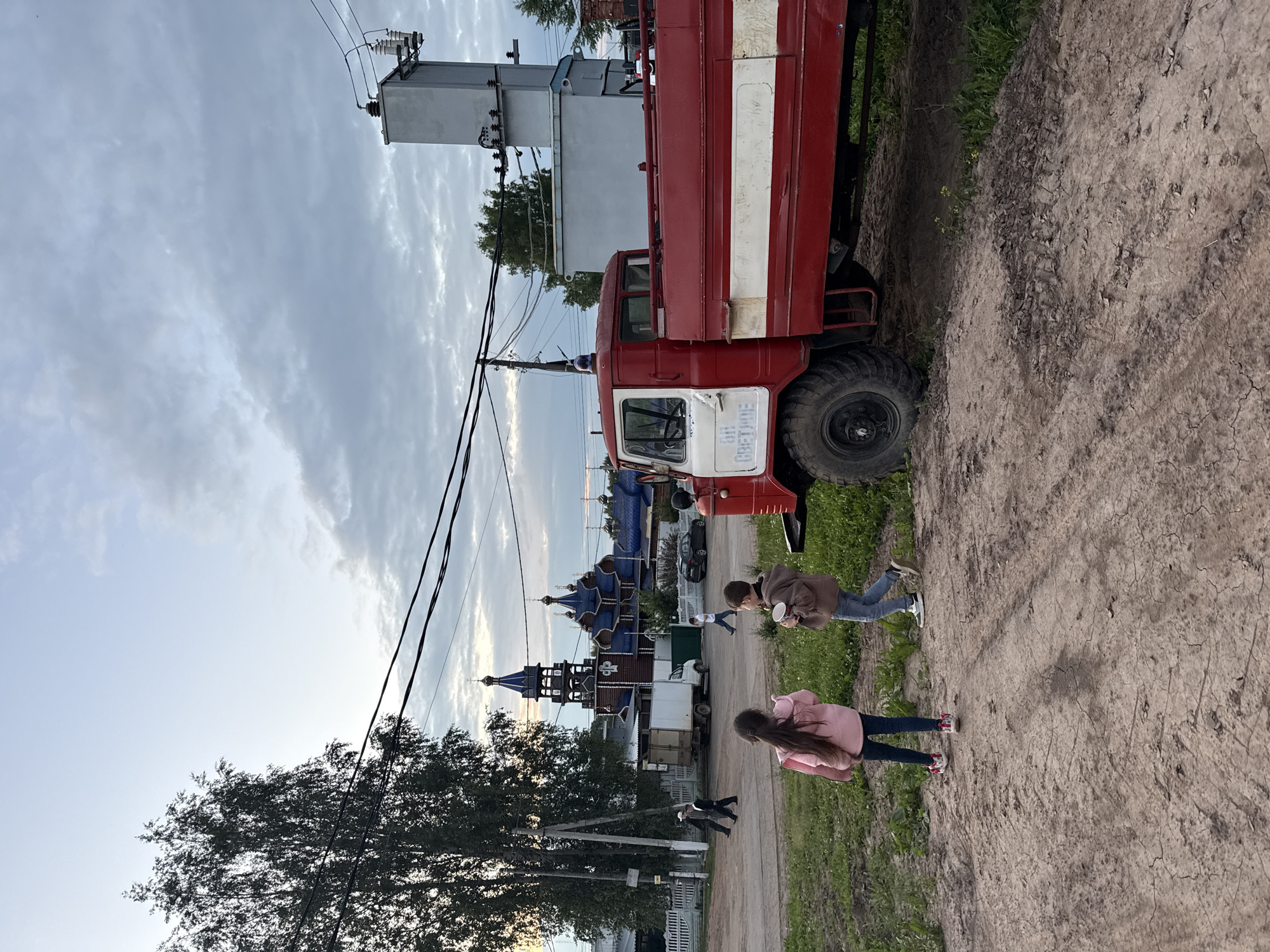 In the village of Kukui, Russia, an outdoor scene unfolds at dusk. The sky is overcast with gray clouds. A large, red and white fire truck, its blue light illuminated, is parked on a dirt road. The truck has a large tire and the text "6H CВETHOE" is visible on its side. Two children, a girl in a pink jacket and a boy in a brown hoodie, stand on the dirt road facing away from the camera. The girl holds a white object, and the boy is eating something from a cup. In the background, several people are walking near a white fence and a white truck. Beyond them, a wooden church with blue domed roofs and a tall bell tower is visible. A power transformer and utility poles with wires are also present in the background, along with a dark-colored car parked near the church. The ground is covered with patchy green grass and dry, cracked dirt.