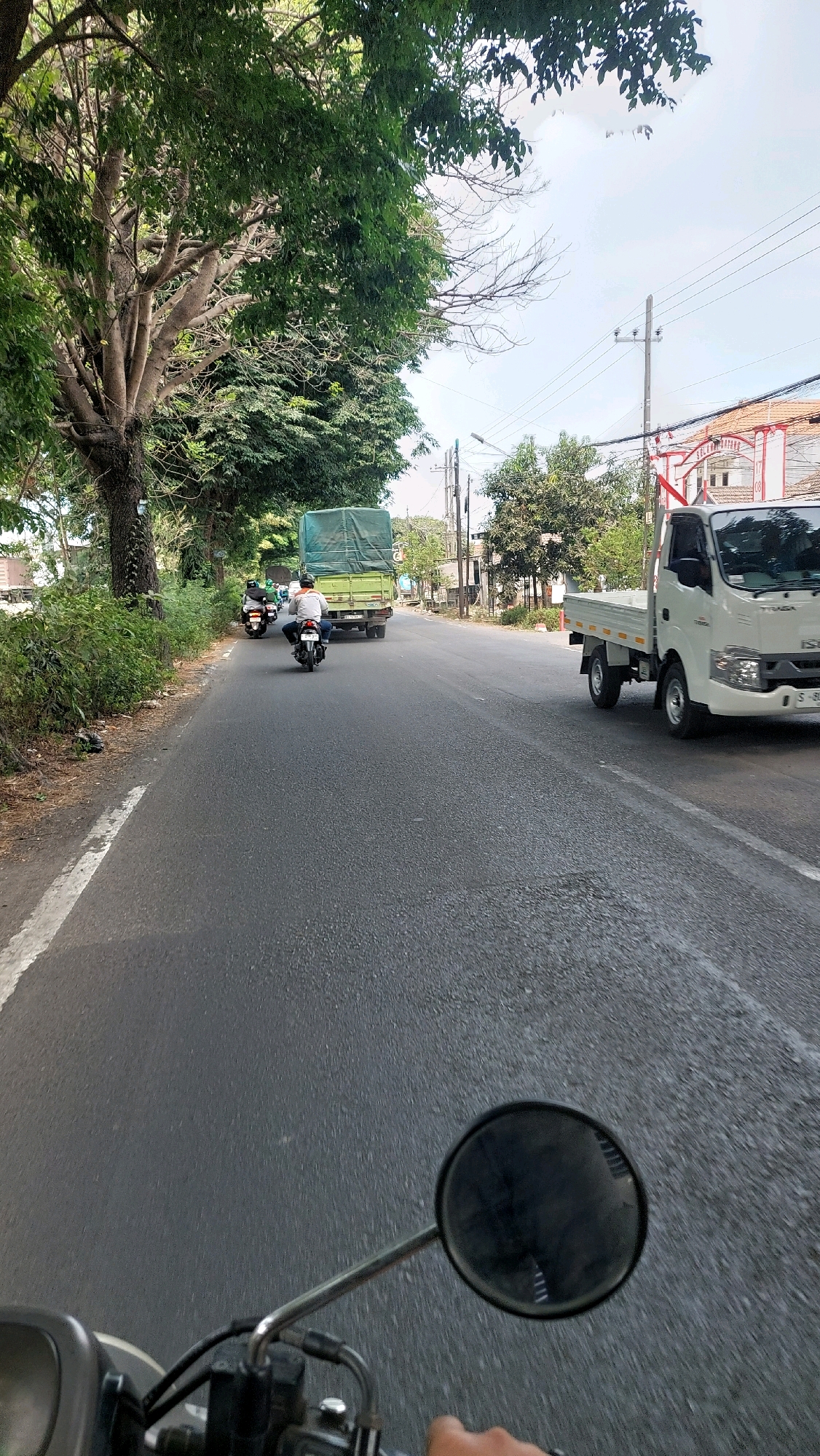 A first-person perspective shot from a motorcycle shows a road in Pilang, Indonesia, with vehicles and greenery. The motorcycle is on the right side of the road, indicated by the handlebar and mirror in the foreground. Ahead, a white flatbed truck with a license plate ending in "SL" is driving in the same direction, on the right lane. Further down the road, a green tarp-covered truck is also traveling. Two motorcycles with riders are visible in the left lane, one is white and the other is black. The road is lined with trees and bushes on the left side, and buildings are visible in the background on the right. The sky is overcast with some clouds.