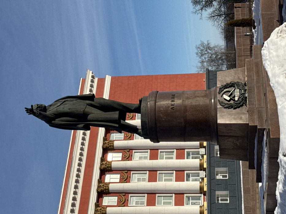 The image displays a bronze statue of Vladimir Lenin situated in Sovietskaya Square, Kursk, Russia, directly in front of the Kursk Regional Administration building. The statue depicts Lenin standing, facing slightly right, with his right arm raised and extended forward, index finger pointed. He is attired in a suit with a long, draped coat. The statue stands on a multi-tiered pedestal of polished reddish-brown granite. The upper cylindrical portion of the pedestal is inscribed with the Cyrillic text "ЛЕНИН" (LENIN). A bronze relief emblem featuring a hammer and sickle surrounded by a wreath is affixed to the lower, broader section of the pedestal.
The Kursk Regional Administration building, a grand multi-story structure, forms the background. Its facade is primarily red, accented by white cornices and numerous white-framed rectangular windows. A central colonnade of white fluted columns with ornate golden capitals dominates a section of the building, with red star motifs visible below some windows. The building's lower section appears to be dark grey.
The scene is under a clear, bright blue sky with faint wisps of clouds, indicating daytime. Patches of snow are present on the ground around the base of the monument and in the foreground, signifying winter conditions. Bare deciduous trees and a small evergreen shrub are visible in the distant background to the right. No people are depicted in the frame.
