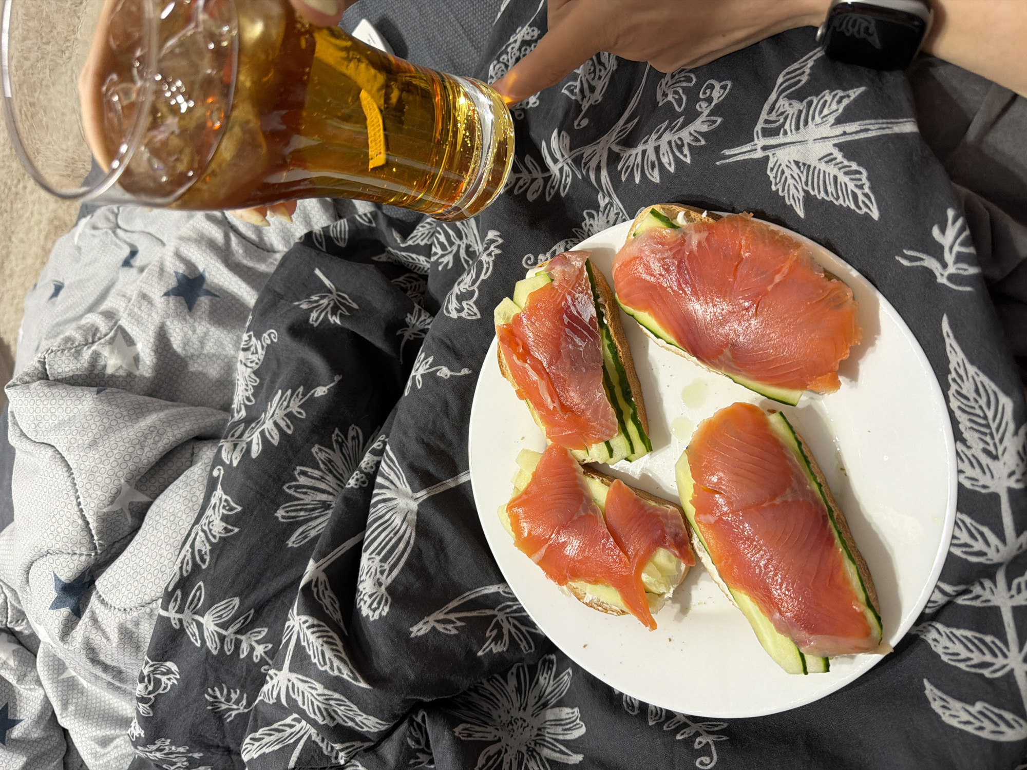 The image presents a top-down view of a relaxed moment, likely indoors on a bed, situated in Chaikovsky, Russia. A person's hand, adorned with a black smartwatch, holds a clear glass filled with a golden-amber drink, complete with ice cubes and a visible lemon slice. The drink appears to be a beverage like beer or iced tea, held at an angle as if about to be consumed.
Adjacent to the drink, on a dark grey blanket patterned with white botanical designs (leaves and flowers), rests a white ceramic plate. On the plate are four open-faced sandwiches or canapés. Each consists of a base, possibly bread or a cracker, spread with a white creamy layer, topped with fresh green cucumber slices, and a generous portion of bright orange smoked salmon. Small drops of oil are also visible on the plate, suggesting a light dressing on the food.
The bedding consists of two distinct patterns: the dark grey floral pattern beneath the plate and the hand, and a lighter grey or white duvet with small dots and larger dark blue stars visible on the left side of the frame. The overall scene suggests a casual meal or snack being enjoyed in comfort, possibly during leisure time. No visible text is present in the image. The setting implies a relaxed and cozy atmosphere.