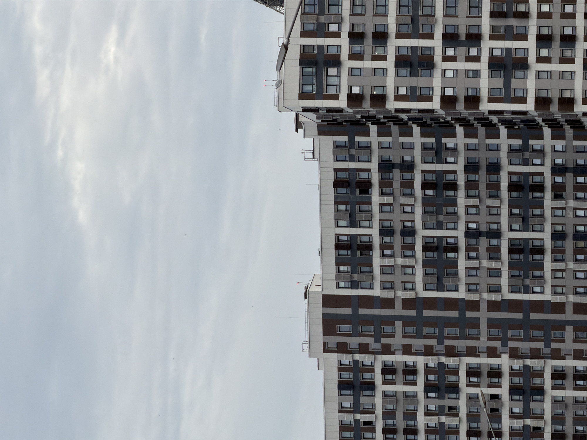 The image shows a modern, multi-story residential building with a facade featuring a geometric pattern of dark brown, gray, and white panels. Numerous windows with closed shutters are visible across the building's exterior. The sky above is overcast with light gray clouds. The building appears to be part of a larger urban development, with hints of other structures and a lamppost in the foreground, suggesting a location in a city. This building is located in the city of Khimki, Russia.