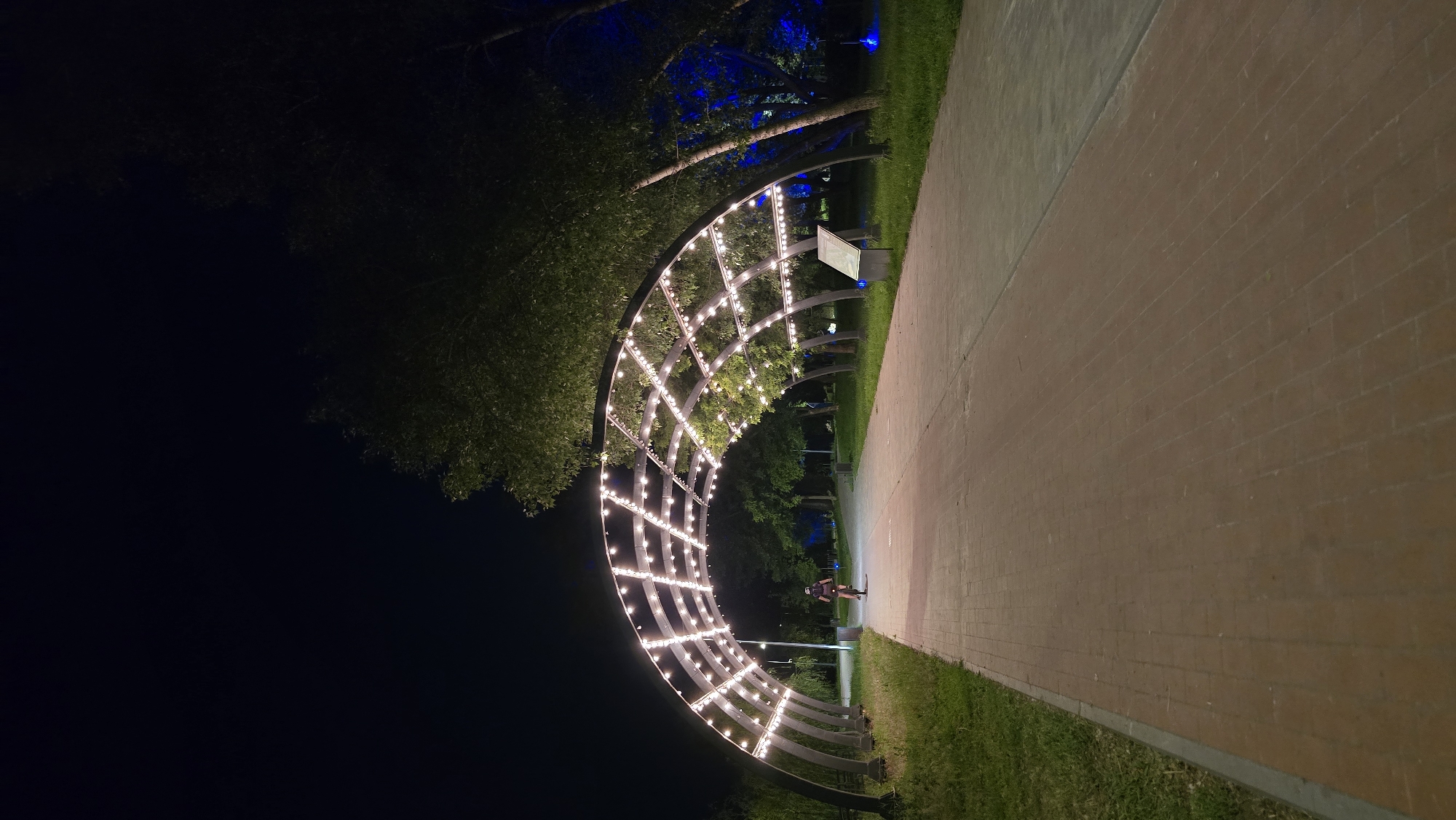 This nighttime vertical shot captures a paved pathway in a park or recreational area, under a decorative lighted archway. The location is an unknown city. The pathway, composed of red brick pavers transitioning to a lighter concrete or stone material further down, extends from the bottom of the frame towards the distance. On the left side of the pathway, a strip of green grass is visible, while on the right, grass and various trees line the path.
A prominent feature is a large, semicircular archway structure spanning the pathway. This archway is constructed from dark metal beams arranged in a grid pattern and is extensively adorned with numerous small, bright white lights, illuminating the path beneath.
Mid-ground, a single individual is visible, riding a bicycle away from the camera, directly beneath the illuminated archway. The cyclist is wearing a dark top and appears to have a backpack.
Further down the path, beyond the archway, the path continues into an area with more trees. One or more streetlights are visible in the distance. To the right of the path, beneath the archway, stands an informational sign or display board with a dark frame and a light-colored surface, angled towards the viewer. Several trees to the right, beyond the archway, are distinctly lit with a vibrant blue light, creating a color contrast with the warm white lights of the archway. The sky overhead is dark, indicating night.