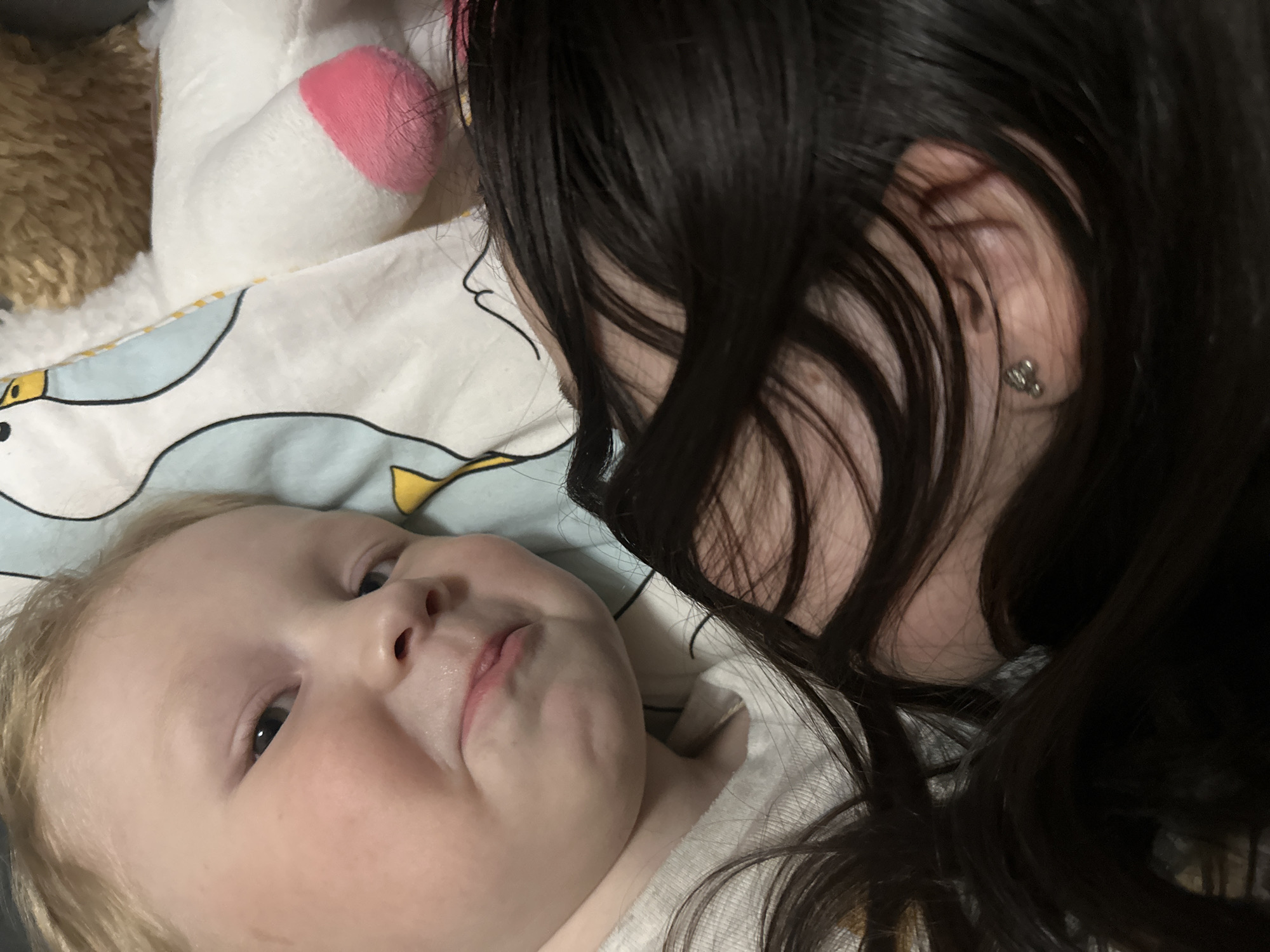 A close-up shot shows a baby lying on a patterned blanket, facing the camera with a slightly puckered expression. The baby has fair skin, blonde hair, and dark eyes. To the right of the baby is a woman with long, dark hair that partly obscures her face. Only a section of her face, including an ear adorned with an earring, is visible as she leans in close to the baby. In the background, to the left, a stuffed toy is partially visible on the blanket. The image appears to have been taken indoors. There is no evidence to suggest that the image was taken in Votkinsk, Russia.