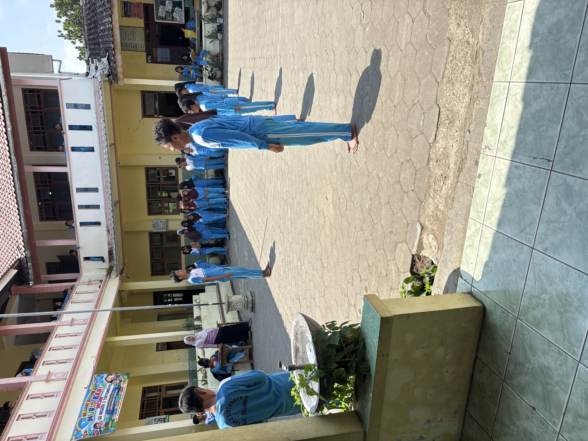 A group of students in matching blue uniforms are standing in a courtyard in front of a school building. The school building has yellow walls and a red tile roof. There are two stories with balconies. A banner on the left side of the building says "SELAMAT DATANG MURID BARU SD NEGERI 2 TIRTOMOYO" which translates to "WELCOME NEW STUDENTS SD NEGERI 2 TIRTOMOYO". The students are standing in rows, facing towards the right side of the image. Some students are looking towards the right, while others are looking forward. The ground is paved with hexagonal tiles. In the foreground, there is a stone fountain with some plants growing around it. The sun is shining brightly, casting shadows on the ground. The overall scene appears to be an outdoor assembly or ceremony at a primary school in Tirtomoyo, Indonesia.
