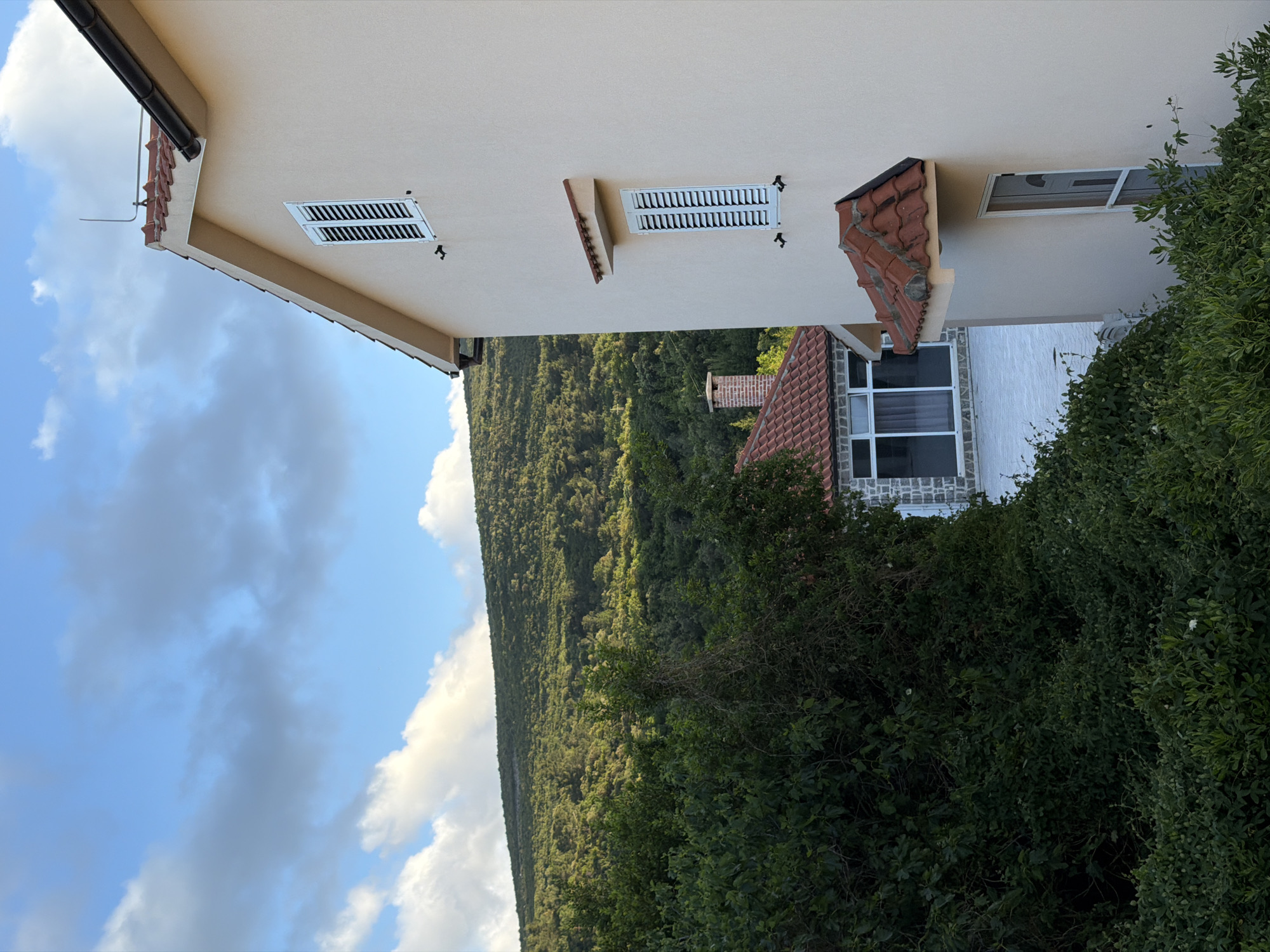 The image shows a view in Gošići, Montenegro, featuring a building with light-colored exterior walls and shuttered windows. The building has a sloping roof with red tiles. The foreground is dominated by green foliage and dense bushes. In the background, there's a tree-covered hillside under a sky with scattered clouds.
