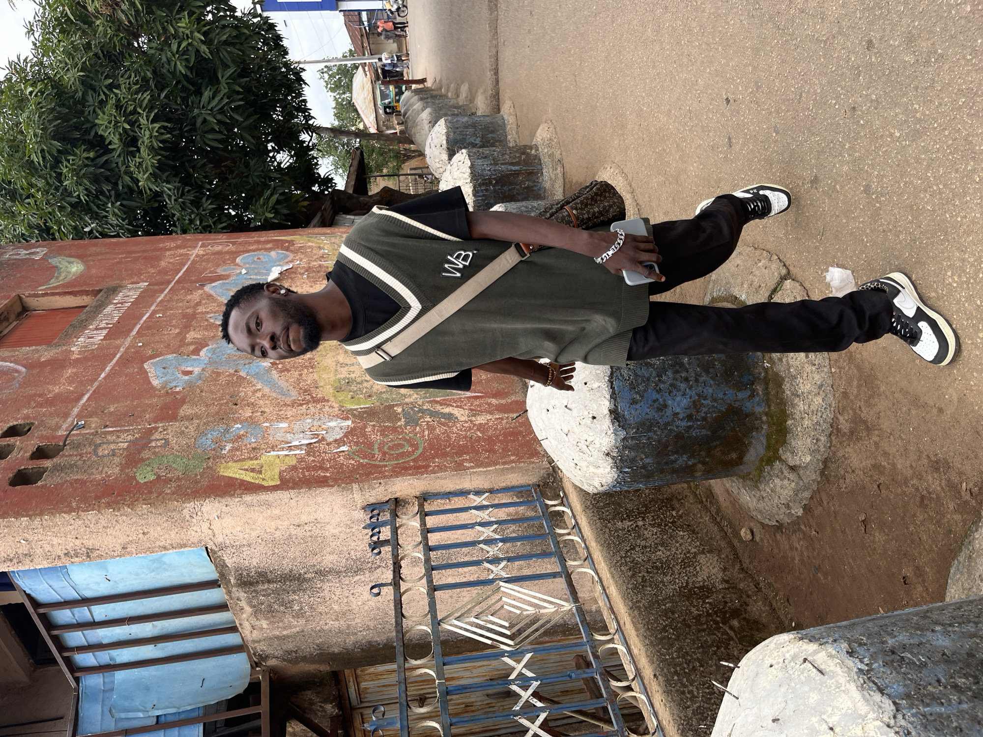 An adult male is depicted walking on an unpaved pathway in an urban area of Jos, Nigeria. He wears a black t-shirt, an olive green V-neck sweater vest with white and black striped trim, black trousers, and black and white sneakers with leopard print detailing on the heels. A tan cross-body bag with a repeating pattern is slung over his right shoulder. He holds a silver smartphone in his right hand and wears a silver cuban link chain bracelet on his right wrist and a brown beaded bracelet on his left. His gaze is directed slightly upwards and to his left.
To the left of the pathway, a weathered, reddish-brown building with faded graffiti, including numeric characters '2' and '4', rises behind a blue-painted metal fence featuring white diamond and X-shaped patterns. An upper section of the building includes a balcony or window secured with blue corrugated metal panels. To the right, a series of aged concrete bollards, some partially painted blue and white, line the path, with a large green-leafed tree situated behind them. In the background, distant buildings, structures, and individuals are visible under an overcast sky.