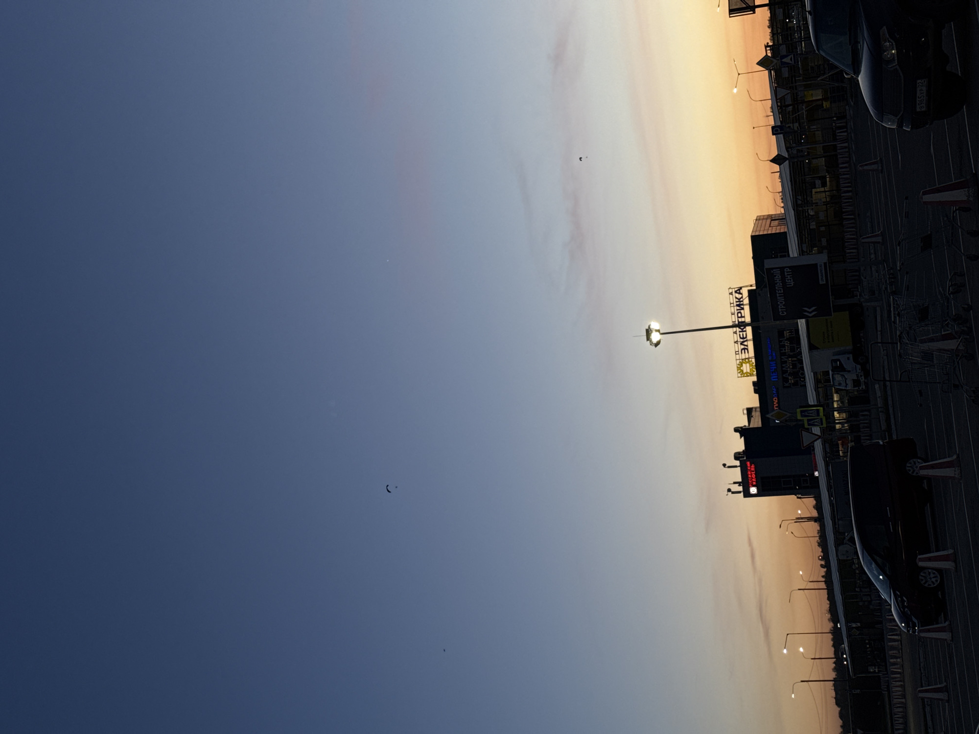 The image, when viewed in its intended landscape orientation, depicts an outdoor commercial or retail area during either dawn or dusk. The sky, which dominates the upper two-thirds of the frame, displays a gradual color transition from a deep, muted blue on the far left to a warm, soft orange and yellow hue towards the right, suggesting the low angle of the sun. A few faint, scattered clouds are visible, and several small, dark bird silhouettes can be seen against the sky, one distinctly crescent-shaped.
The lower portion of the image reveals a dark, asphalt surface, likely a parking lot, with several vehicles partially visible along the right side. One vehicle on the far right has a license plate reading "B 945 XP 23", indicating a Russian location, specifically Krasnodar Krai. A cluster of shopping carts and several white and red striped barriers or traffic cones are also present.
Along the horizon line, several commercial buildings are discernible, appearing as dark silhouettes against the glowing sky. A tall, brightly lit streetlight stands prominently near the center-right. Several other streetlights are visible further in the background. Prominent Cyrillic signage on the buildings includes "ЭЛЕКТРИКА" (Elektrika, meaning Electricity/Electronics) and "СТРОИТЕЛЬНЫЙ ЦЕНТР" (Stroitel'nyy Tsentr, meaning Construction Center). Another sign appears to reference "РЕСТОРАН И КАФЕ" (Restoran i Kafe, meaning Restaurant and Cafe). Various smaller traffic or directional signs are also scattered throughout the scene.
No people are visible, and no specific activities are taking place, contributing to a calm and serene atmosphere. The scene captures the quiet transition period of a commercial zone as day turns to night or vice-versa.