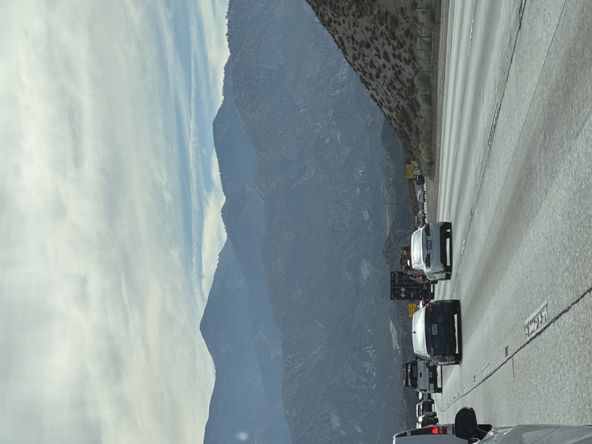 The image captures a traffic scene on a highway in Oak Hills, United States. In the foreground, a white car is partially visible on the left, its side mirror prominent. The highway stretches ahead, with several vehicles in motion. A dark blue sedan is in the middle lane, followed by a white sedan in the right lane. Beyond them, a white pickup truck and a large black trailer are also on the highway. The environment is mountainous, with layered, blue-gray mountain ranges dominating the background under a partly cloudy sky. The slopes of the mountains are covered with dry brush and sparse vegetation. Road signs are visible on the right side of the highway, indicating some services or warnings. The overall scene depicts a typical highway commute in a mountainous region.