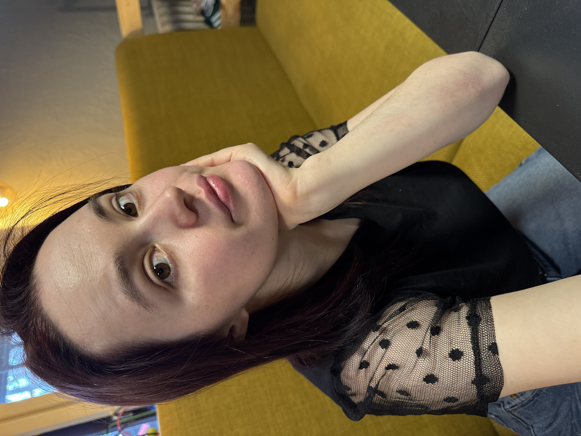 A woman with dark hair and fair skin is resting her head on her hand while sitting on a yellow couch. She is wearing a black shirt with polka dot mesh sleeves. Her hair has a reddish tint in the light. She is sitting at a black table, wearing blue jeans. The image shows an interior, probably of a cafe or lounge, although the specific location in Izhevsk, Russia cannot be determined from the provided visual information.