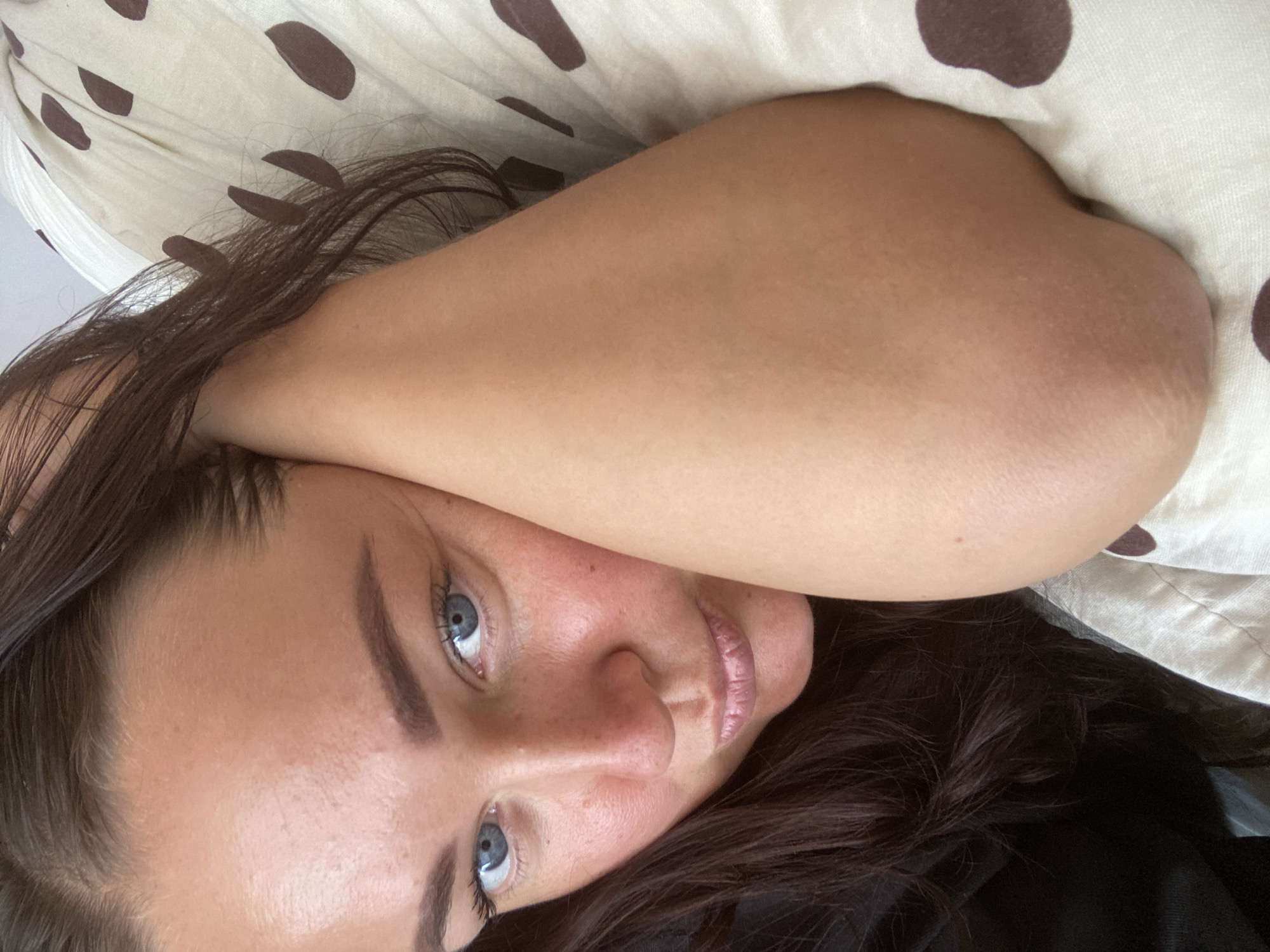 A close-up, high-angle, indoor selfie shows a young woman with dark brown hair and blue eyes resting her head on her arm. She is lying on a bed with white and brown polka dot bedding. The woman is wearing a black top. The lighting is soft, and the focus is on her face. There is no identifiable information to place this image in Sochi, Russia.