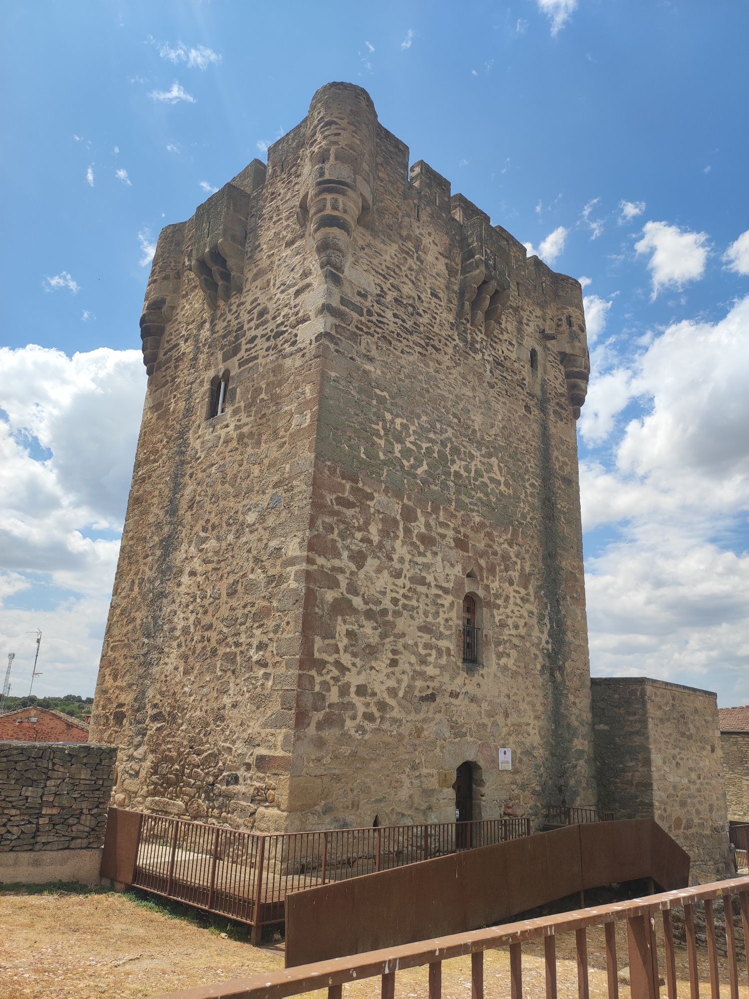 The image shows the Torre de Villamizar in Sobradillo, Spain. The tower is a tall, square stone structure with crenellations at the top. It has small windows and a doorway at the base, with a modern metal ramp leading up to it. The tower is surrounded by a stone wall and dry grass. The sky is blue with scattered white clouds. In the background, there are some trees and buildings. The overall impression is one of a historic landmark in a rural setting.