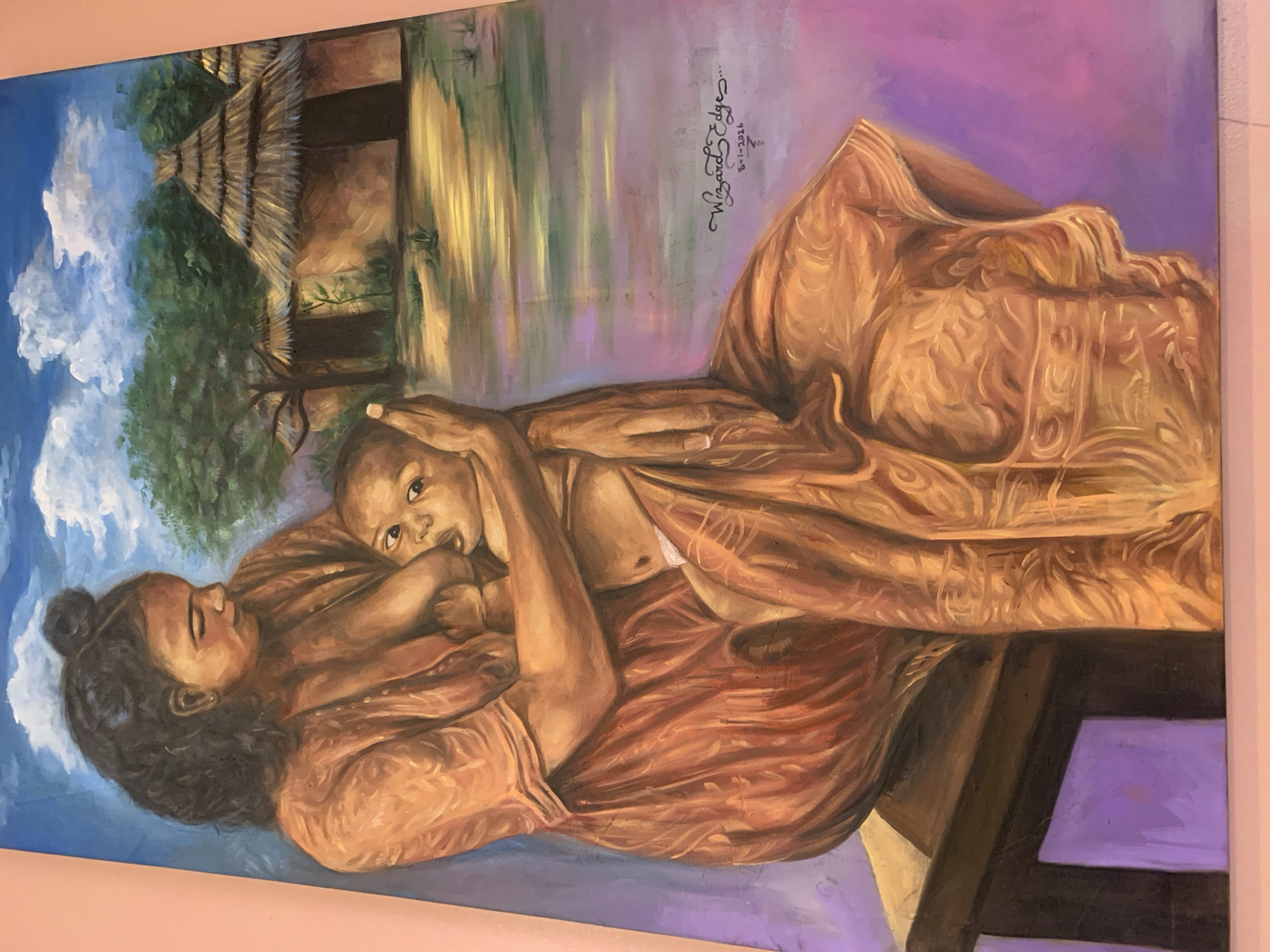 A painting depicts a woman breastfeeding a baby. The woman has dark hair pulled into a bun and is wearing an orange garment. The baby, with wide eyes, is nestled against the woman, also wearing a light-colored garment. They are seated on a wooden bench or stool. In the background, there are traditional thatched-roof huts surrounded by trees under a blue sky with white clouds. The ground in the background appears to be a body of water or wet earth with reflections. A signature and date, "Wizard Edge..." and "8-1-2026", are visible in the lower right portion of the painting. The artwork is displayed against a pink wall. The painting's style suggests it might be a representation of rural life, potentially in a place like Jos, Nigeria, given the architectural style of the huts and the depiction of the people.