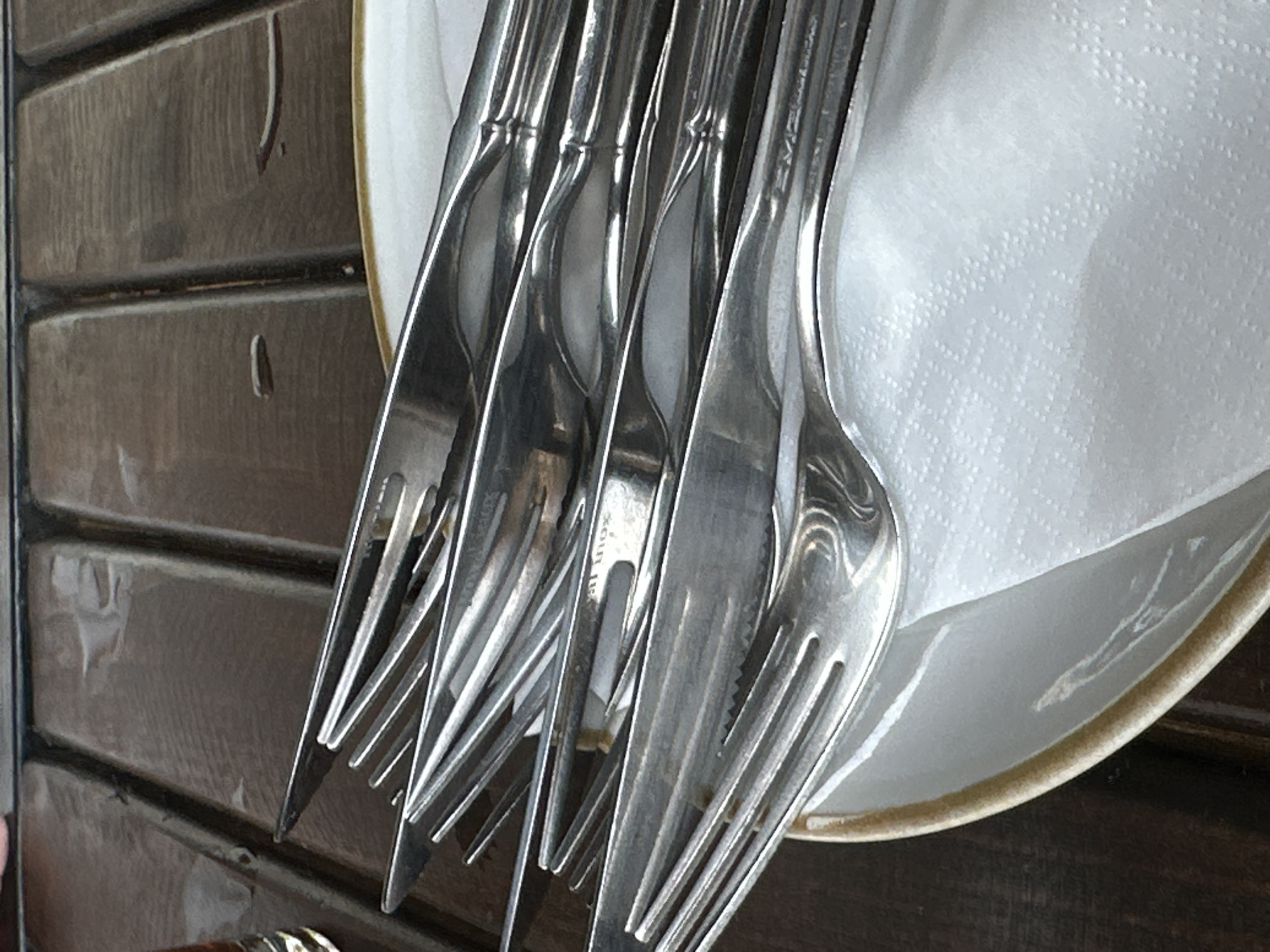 Forks sit atop a white plate with a gold rim, which is likely set on a dark, horizontally-oriented wooden surface. The forks appear to be metal and are bunched together. The surface may be part of an outdoor patio or table at a restaurant in Bar, Montenegro. There is a textured, white cloth on the plate with the forks.