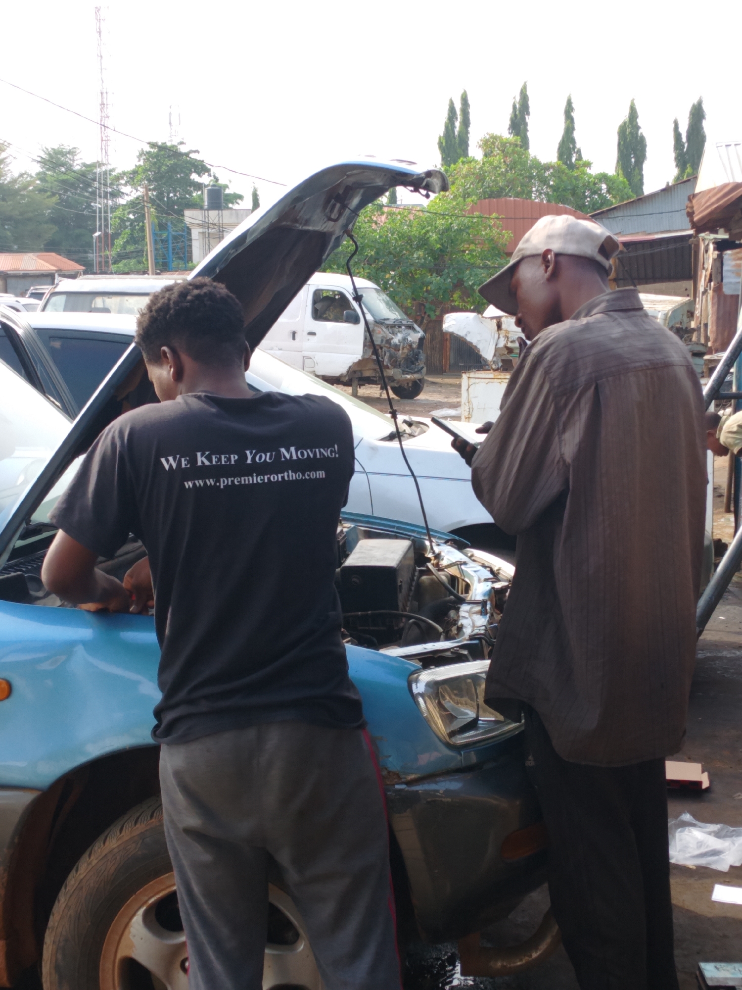 The image depicts two men working on a light blue car with its hood open. The scene is likely in Kakuri Gwari, Nigeria.
One man, facing the car, is wearing a black T-shirt with white lettering that reads "WE KEEP YOU MOVING! www.premierortho.com," and gray sweatpants. He appears to be examining or working on the car's engine. The other man, standing to the side and facing away from the camera, wears a tan baseball cap and a brown button-down shirt and black pants. He holds a smartphone in his hand.
The car is parked outdoors. There are other vehicles visible in the background, including a white van with front-end damage. A tall metal structure with antennas is visible in the distance. There is a simple, functional atmosphere suggesting an auto repair shop or a similar business. The environment appears urban, and the lighting suggests it is daytime.