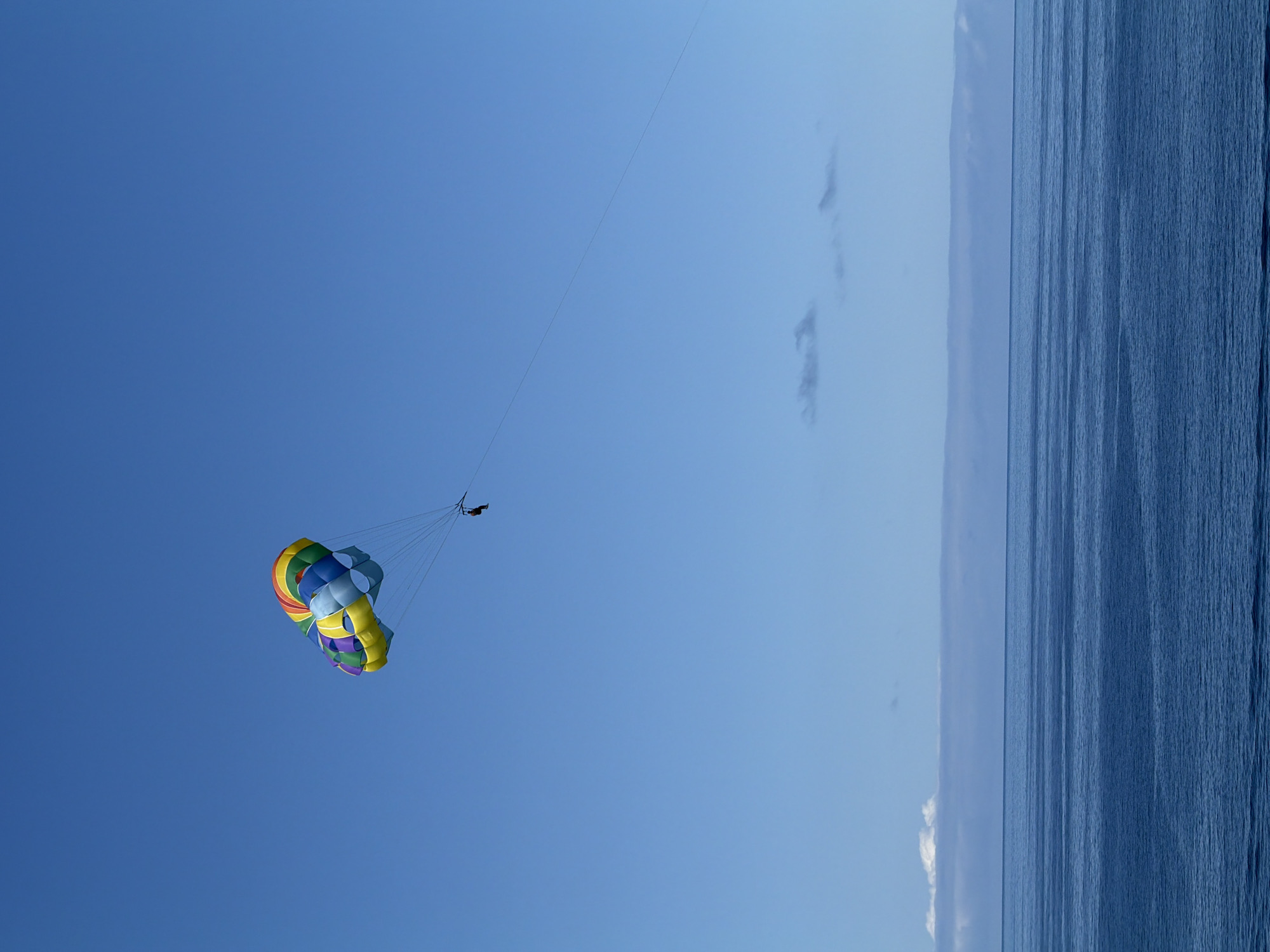 The image depicts a parasailer in mid-air over the ocean. The parasail is multicolored, with blue, yellow, green, and red sections. A thin tow rope extends from the parasail towards the upper right. Below the parasailer is the ocean. The sky is clear. This activity is likely taking place near the city of Funchal, Portugal.