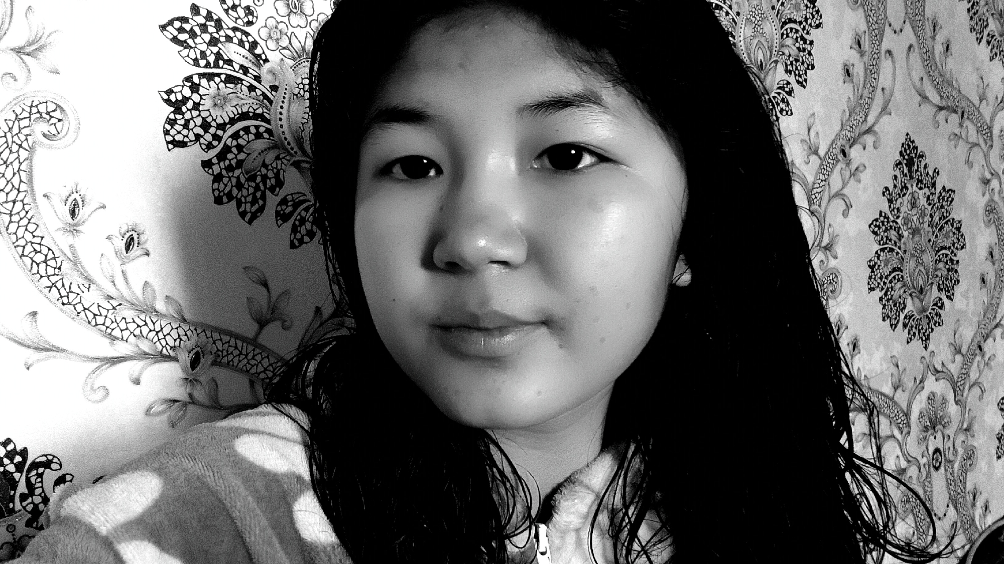 A black and white, medium close-up selfie shows a young East Asian female facing the camera. She has long, dark, wet hair that falls around her shoulders and part of her face. Her skin is smooth with some blemishes visible. Her eyes are dark and her lips are slightly parted. She is wearing a light-colored, zip-up garment, possibly a hoodie or jacket. Behind her, the wall is covered in a dark, floral wallpaper with a repeating pattern. The lighting creates shadows on the left side of her face and on the garment she is wearing. The overall scene appears to be indoors. The location of Uchkun, Kyrgyzstan is not depicted or inferable from the image.