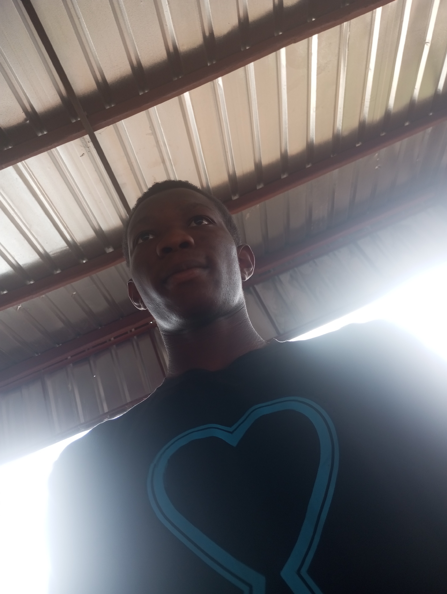 A low-angle, slightly distorted shot shows a young Black male from the chest up, looking up and to the right. He is wearing a black t-shirt with a light blue outline of a heart design on it. The background consists of a corrugated metal roof with visible wooden beams, suggesting an outdoor or semi-covered structure, possibly a workshop or market stall. Bright sunlight streams in from the right side of the frame, creating a strong glare. The image appears to be taken in Onitsha, Nigeria.