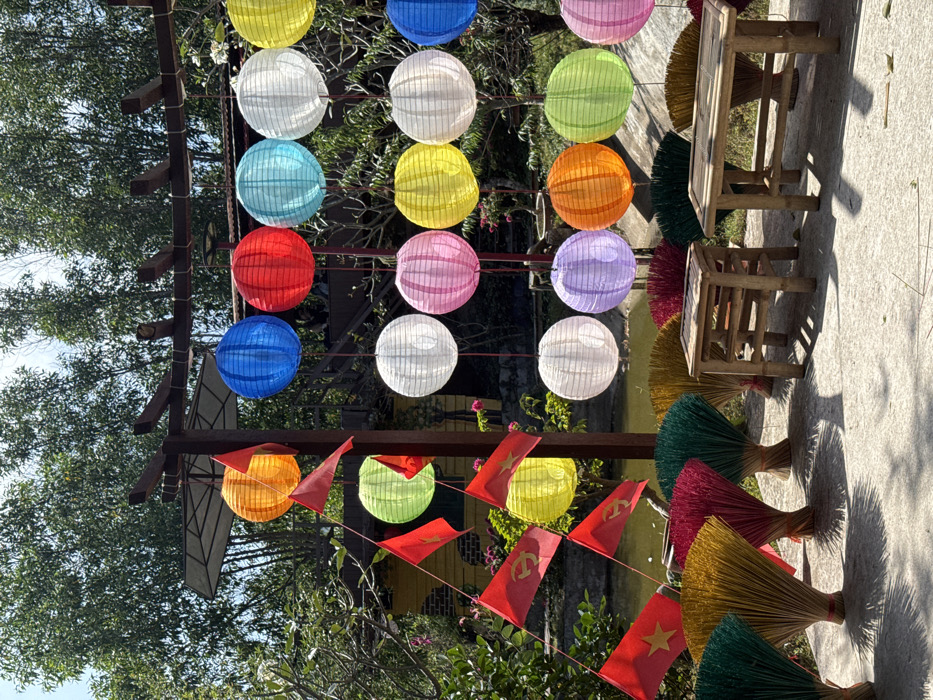 A vibrant display of colorful lanterns hangs from a wooden structure amidst lush greenery. Red flags with gold stars flutter below. Beside the lanterns, small wooden tables and chairs rest on a concrete surface. The scene is filled with visual textures from the trees, bamboo, flags, and lanterns, creating a lively, festive atmosphere. The overall perspective offers a glimpse into an outdoor setting.