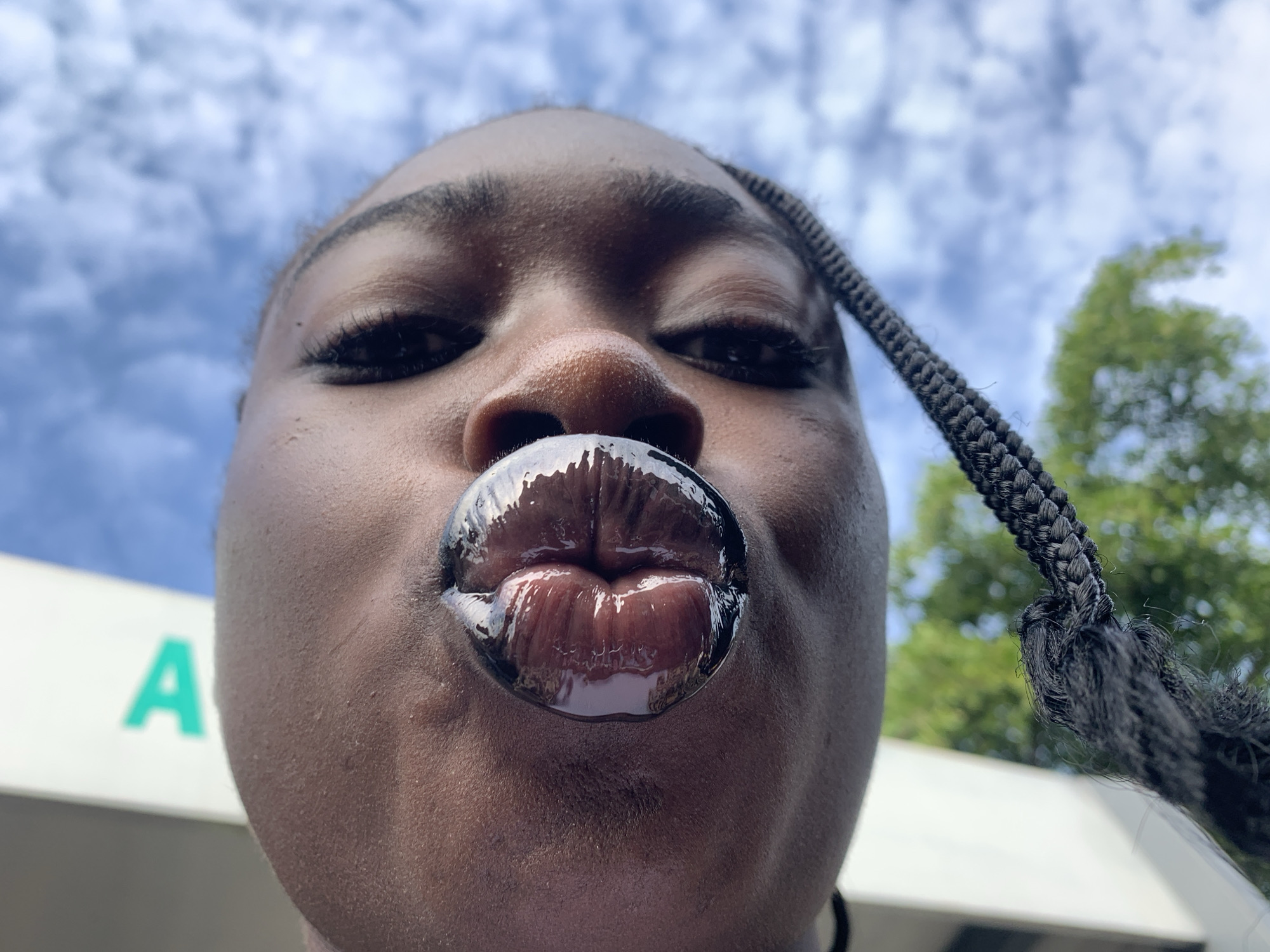 In this image, a close-up, low-angle shot captures a person's face, focusing on their mouth which is pursed and covered in a dark, glossy lip gloss. The person has dark skin and braided hair visible in the frame. Behind them, a bright blue sky with white clouds is seen, along with hints of green foliage and a portion of a white structure with a green letter "A" on it. The composition is vertical, with the person's face tilted upwards and towards the camera. The image appears to be taken outdoors.