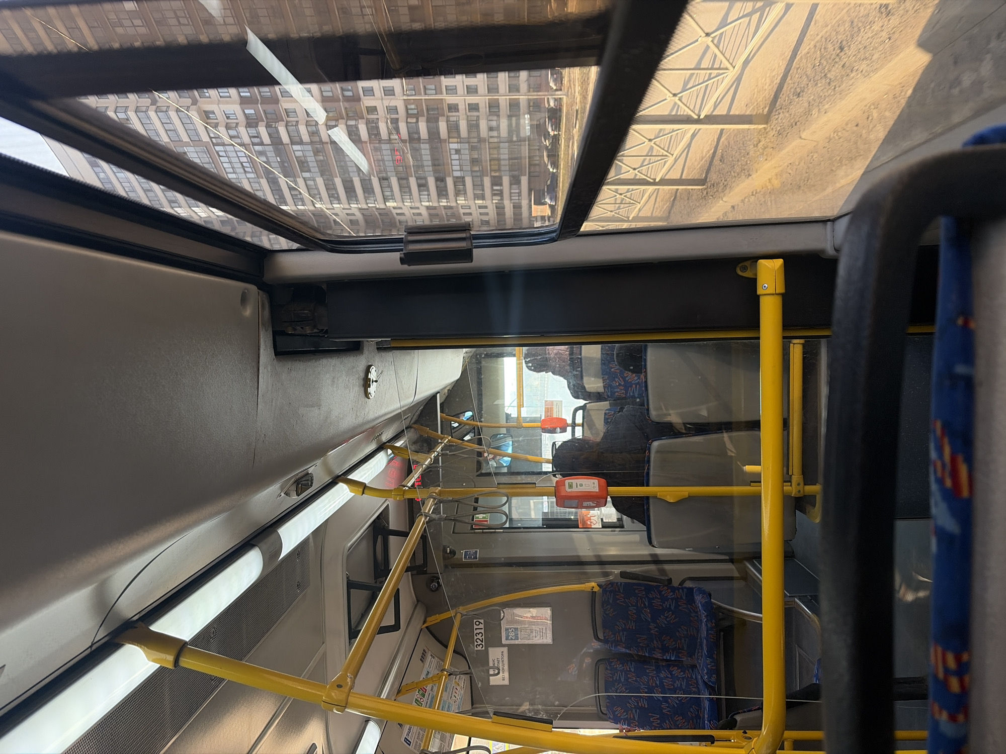 The image depicts the interior of a bus in Saint Petersburg, Russia. Rows of blue patterned seats are visible, separated by yellow metal support bars. Several passengers are seated in the back, while the front of the bus is largely empty. Through the bus windows, a large office building with a repetitive window pattern and a pedestrian overpass outside the bus are visible, suggesting an urban environment. The ceiling of the bus features a skylight showing a reflection of nearby buildings. Several informational stickers and safety notices are posted on the bus walls.