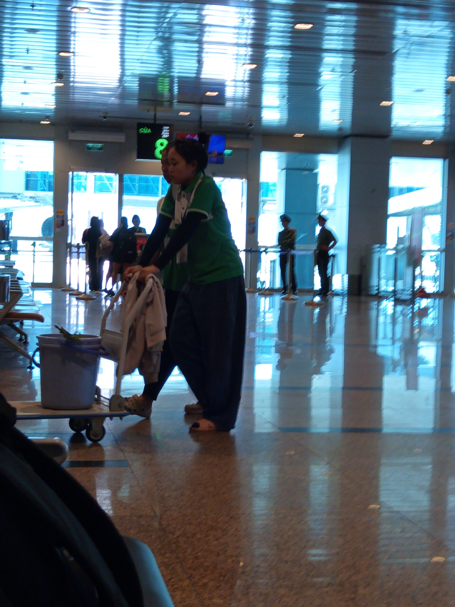 The image depicts a busy interior scene, likely an airport terminal, in Bắc Cam Ranh, Vietnam. Two individuals are the central focus. The person on the right is wearing a green shirt with black sleeves and dark pants, pushing a cleaning cart. This cart has a light-colored bucket and what appears to be folded laundry or cleaning cloths. The person's face is visible, looking down. To their left, another individual, also wearing dark pants and a lighter shirt, is partially visible and appears to be assisting or accompanying the cleaner.
In the background, several other people are present, some standing near what looks like a gate or information display. Two individuals in uniform, possibly security personnel, stand near the glass doors further in the background. The environment is characterized by a highly reflective floor, possibly granite or polished tile, with light reflecting from a ceiling structure made of horizontal panels, suggesting a modern architectural design. The overall lighting is bright, with some areas appearing overexposed due to the reflections. A digital display board showing the number "8" is visible in the upper portion of the image. The scene conveys a sense of ongoing activity, typical of a public transit hub.