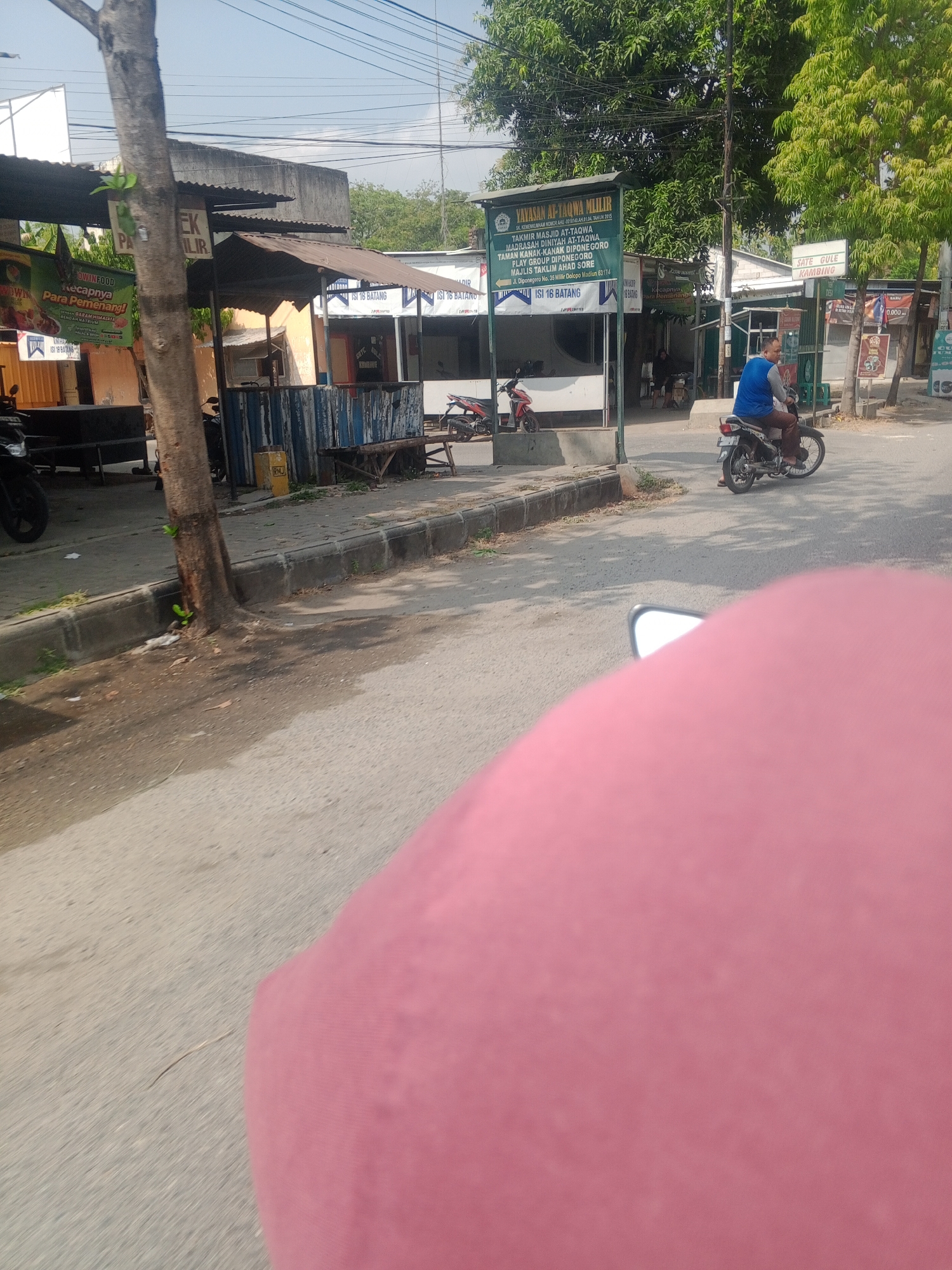 The image captures a street scene in Glonggong, Indonesia. A person on a motorcycle is riding on the road, wearing a blue shirt and brown pants. In the background, there is a small building with a sign that reads "YAYASAN AS-TAUWA MLILIR" and "TAK MIR ILIR". Several other buildings and trees are visible. The day appears to be sunny.