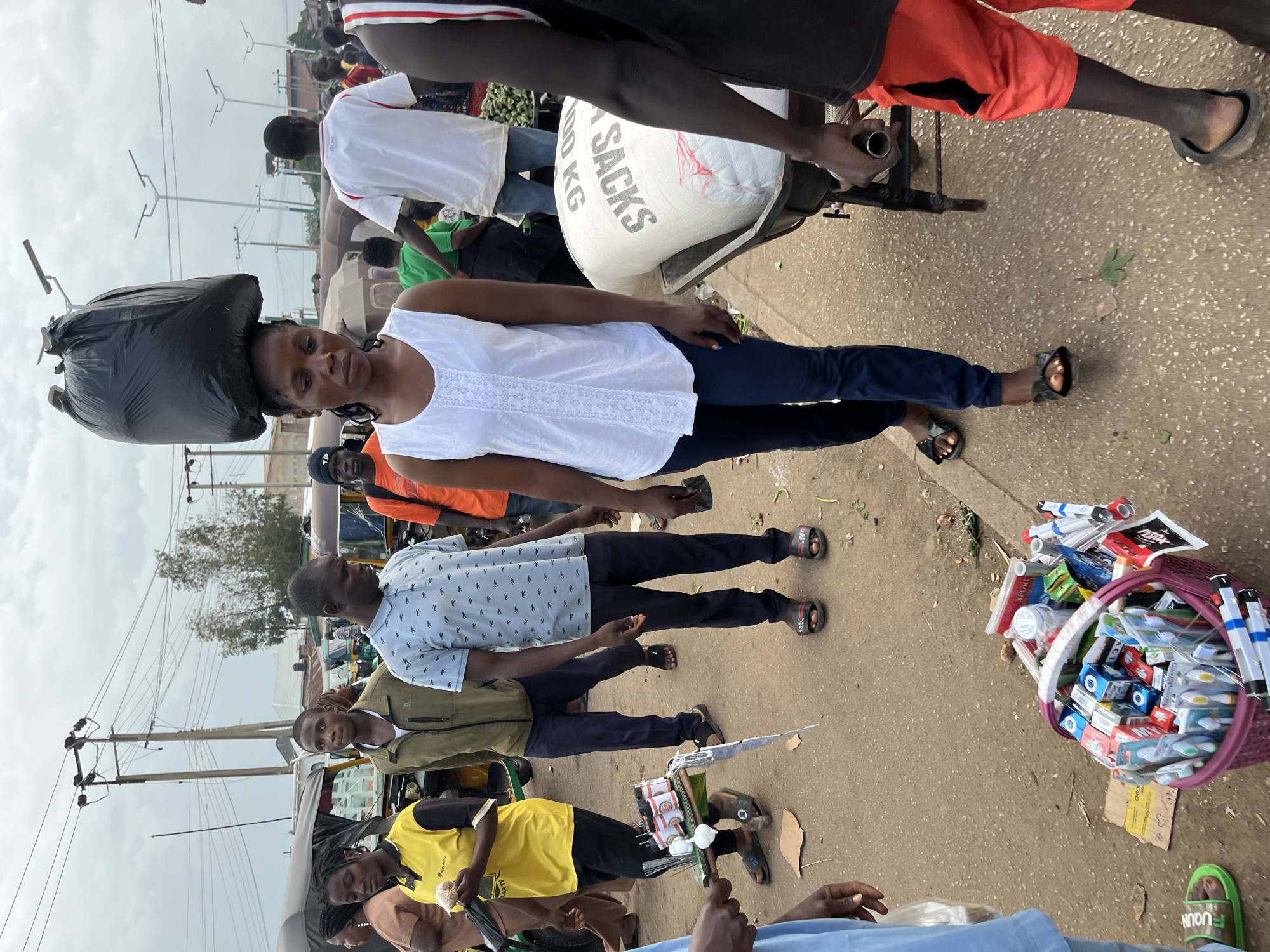 A young woman with dark skin and braided hair balances a large black trash bag on her head while walking forward in a marketplace. She is wearing a white sleeveless top with lace detailing and navy blue pants. She is looking down and slightly to her right. She is in motion, with her right leg forward.
In the background, other people are walking and standing around. To the woman's left, a person in a yellow shirt is holding what appears to be a bag of snacks or food. To her right, a large sack labeled "100 KG" is being loaded onto a cart. A person in a red shirt is visible on the far right of the frame, holding a handle of the cart.
The environment appears to be a dusty outdoor market, with a paved ground and some greenery visible in the distance. There are also overhead power lines and streetlights, indicating an urban or semi-urban setting. The sky is overcast.
The image was captured in Bukuru, Nigeria.