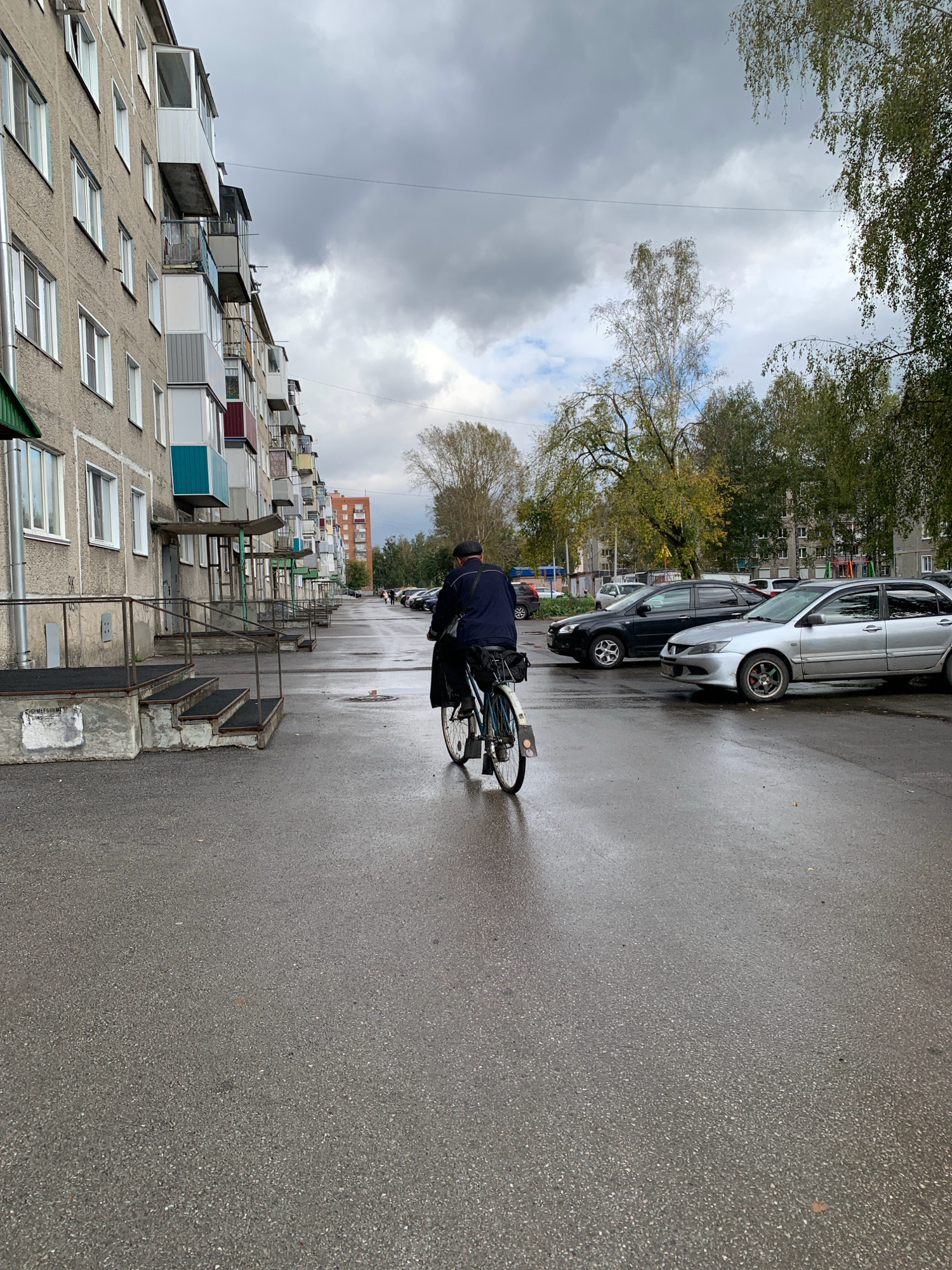 The image depicts a street scene in Prokopyevsk, Russia, characterized by overcast skies and wet asphalt. A person on a bicycle rides away from the viewer down a street lined with multi-story apartment buildings. The buildings, typical of Soviet-era architecture, have a rough concrete facade and balconies. Cars are parked along the right side of the street. Trees with autumn foliage are visible in the background. The overall atmosphere suggests a cool, damp day in an urban environment.