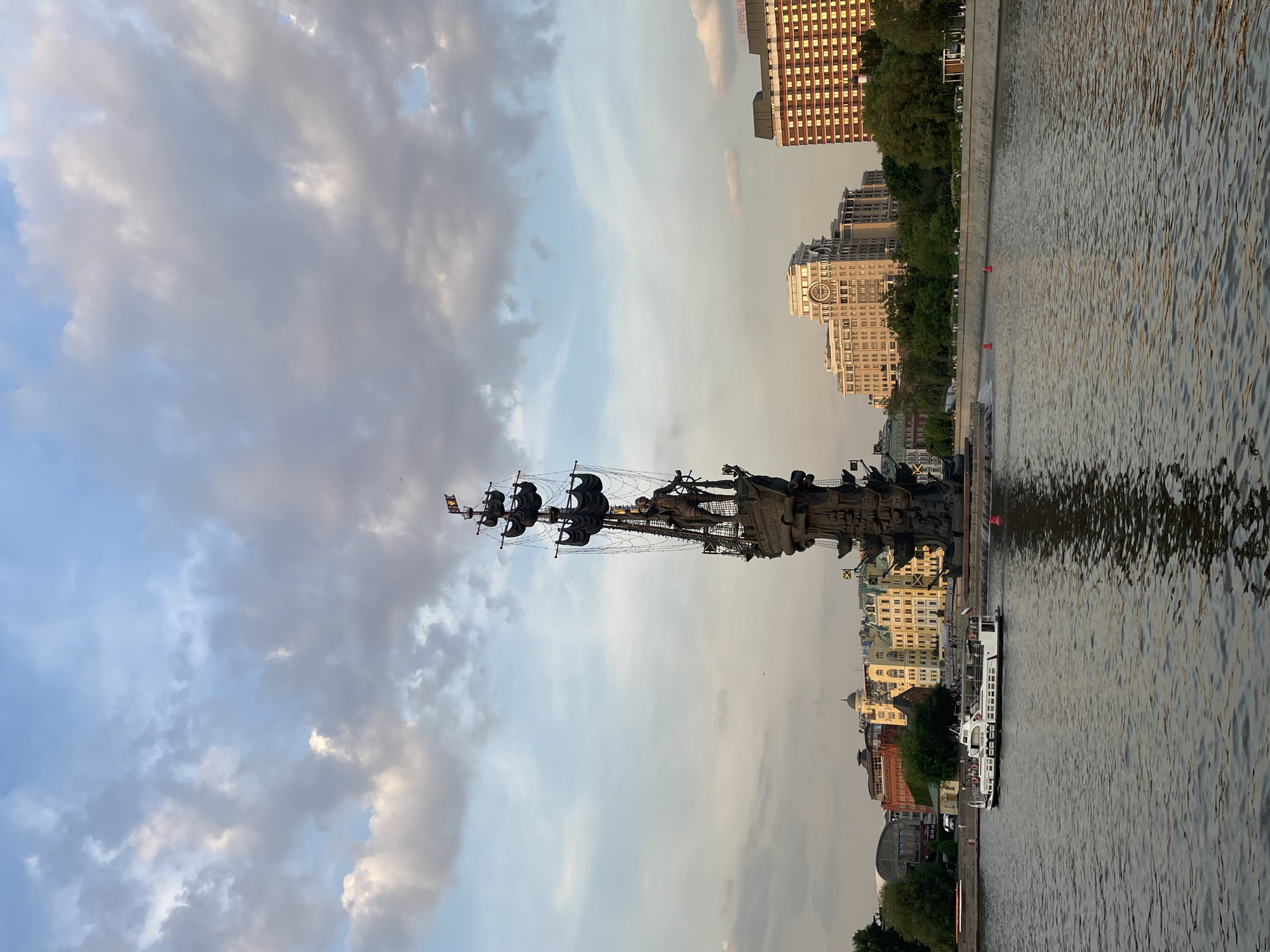 The image shows the Peter the Great Statue in Moscow, Russia, next to the Moskva River. The statue depicts Peter the Great standing on a ship. Several buildings are visible in the background, with a mix of older and modern architecture. The sky is partly cloudy. A small passenger boat is docked near the statue. The river is reflecting the buildings and the statue.