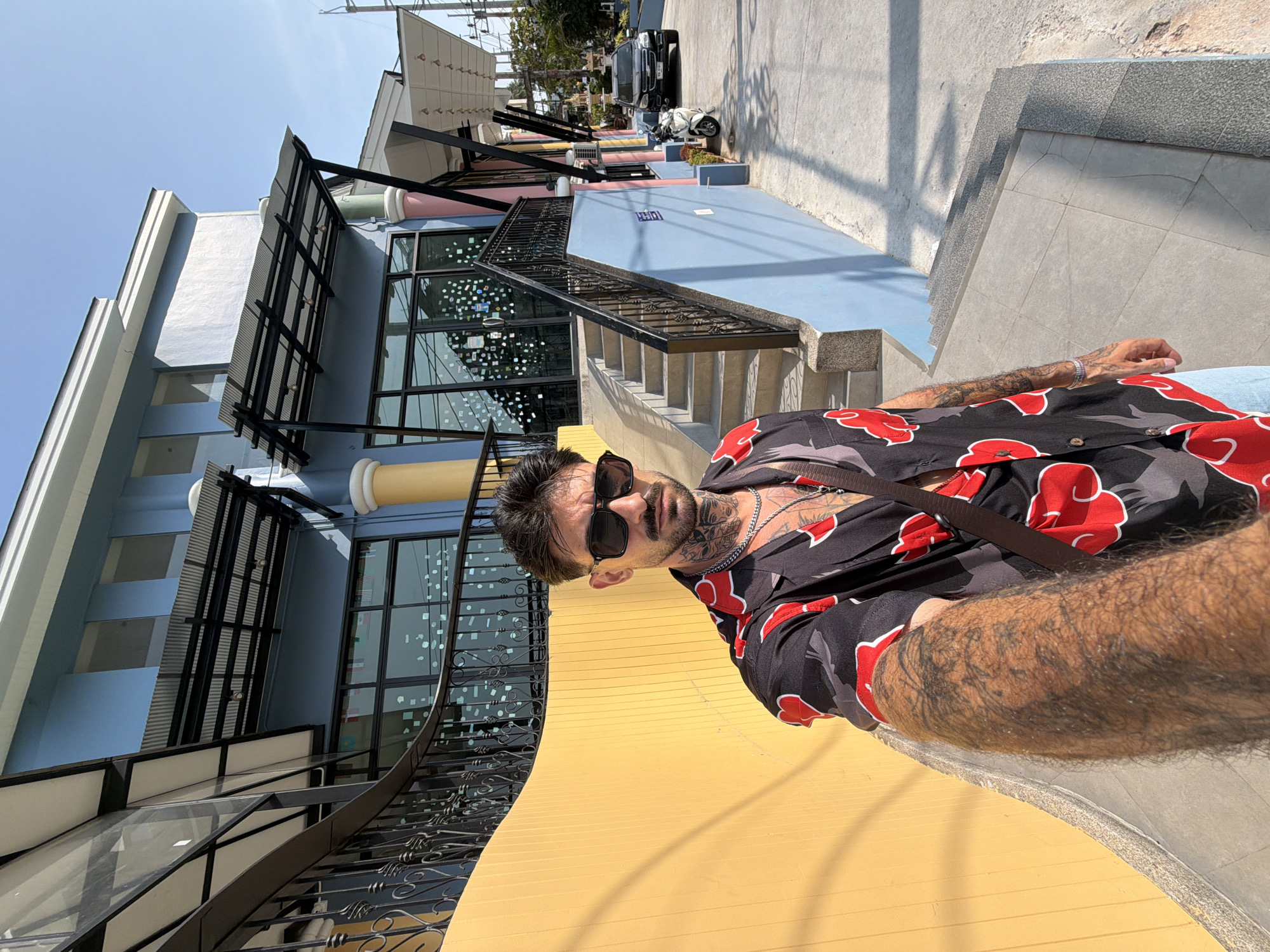 An eye-level outdoor shot, rotated ninety degrees clockwise, features a man posing in front of a colorful building complex, identified as Patong, Thailand. The bright midday sun casts clear shadows, indicating sunny weather.
The primary subject is a man with dark hair, a beard, and numerous tattoos on his arms and neck. He wears black sunglasses, a silver chain necklace, and a black short-sleeved button-up shirt adorned with red cloud patterns, reminiscent of the Akatsuki organization from Naruto. He gazes directly at the camera with a neutral expression. His right arm, heavily tattooed and hairy, rests out of frame, while his left arm is partly visible. He appears to be leaning against or sitting on a vibrant yellow surface in the foreground.
Behind the man, a multi-story building complex with a modern design is visible. The closest section is light blue, featuring large glass windows reflecting the sky and interior details. Black metal railings and staircases with intricate designs ascend alongside the building. Further back, other sections of the buildings display colors like pink, light grey, and white, creating a lively architectural backdrop. In the distance, on what appears to be a street or pathway, a dark car (possibly an SUV) and a white scooter are parked. Small green plants are placed in planters near the street. No discernible text is visible in the image.