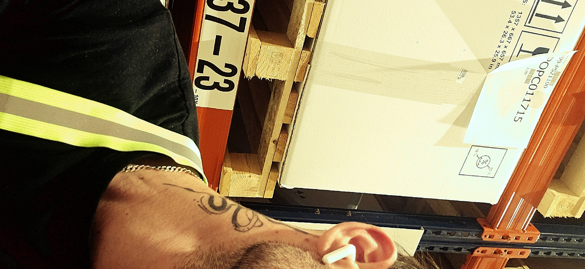 A close-up shot presents a partial view of a person wearing a reflective vest, a gold chain, and an earbud in their ear. The person has a tattoo on their arm. Adjacent to them is a warehouse shelf, filled with items including a wooden pallet. On the pallet, there is a large, white box with shipping labels and handling instructions marked on it. The label has text that reads "899-PS21100", "POPC011715", along with measurements in both metric and imperial units. The environment suggests an industrial or storage setting. There is no location information or additional context to suggest the photo was taken in Plovdiv, Bulgaria.