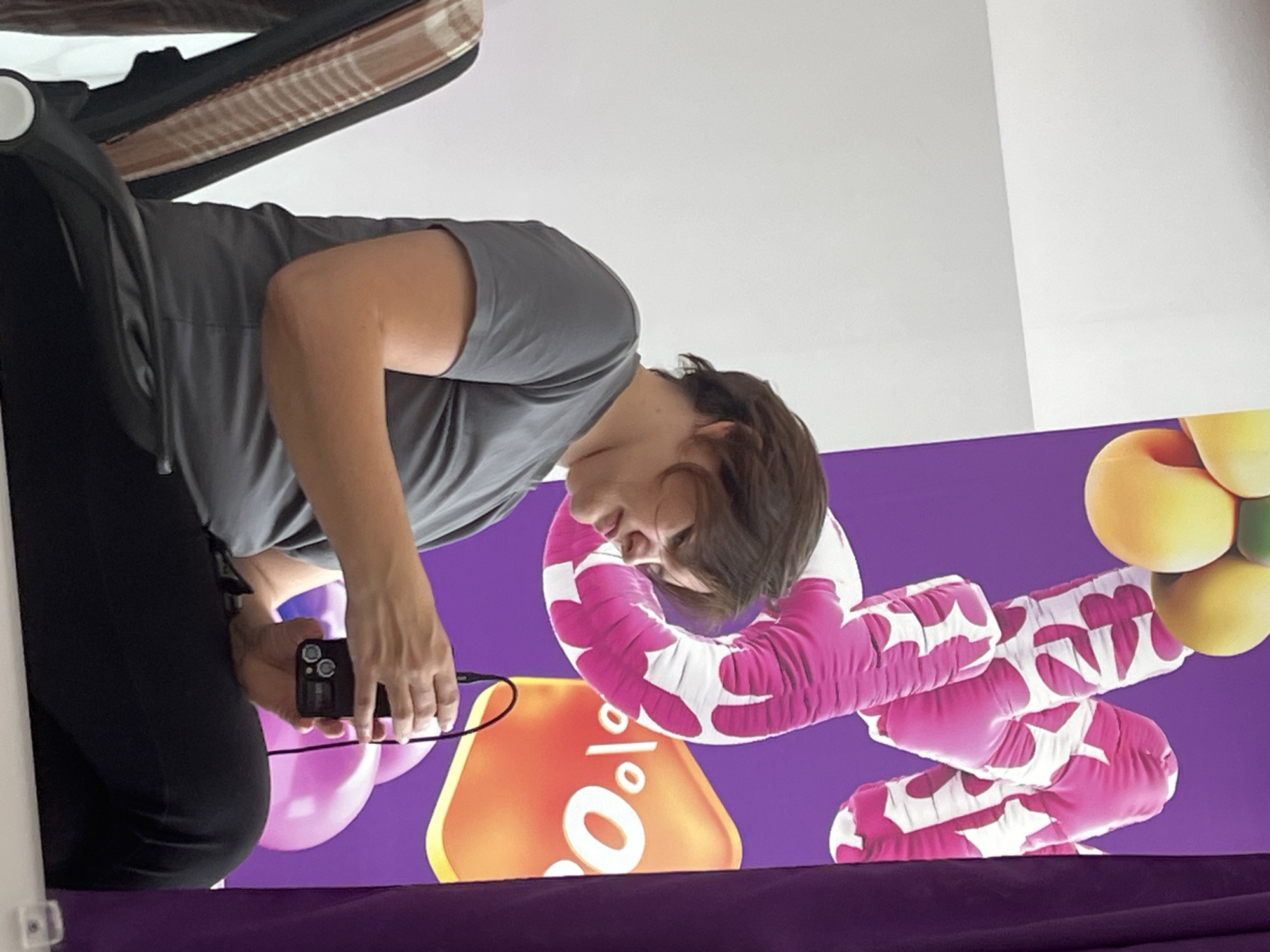 A person is seated in a chair, facing to the right and looking down at a small black electronic device in their hands. The person is wearing a gray t-shirt and black pants. Behind the person, there is a purple banner with colorful, balloon-like graphics and text that includes "o|%". The background to the left is a white wall. The scene appears to be indoors, possibly at an event or exhibition. The location is identified as Afipskiy, Russia.