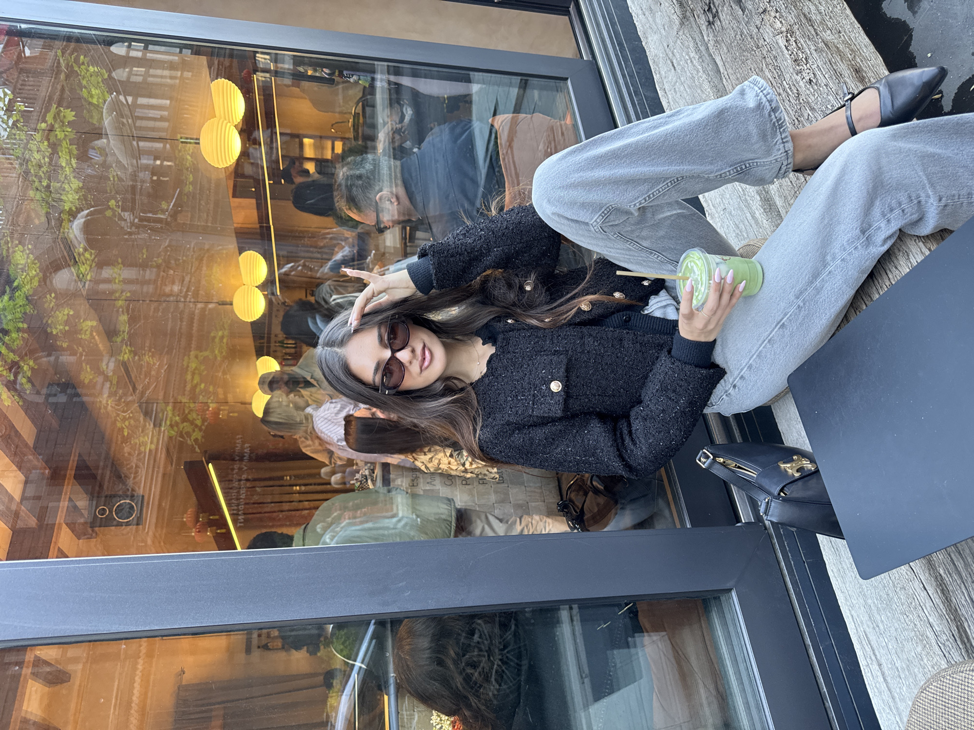 The photograph depicts a young woman sitting at an outdoor table in Yerevan, Armenia. She is wearing a black tweed jacket, light-wash jeans, and black Mary Jane shoes. She is holding a green beverage with a straw in her right hand and posing with her left hand raised near her forehead. She is wearing sunglasses.
A black Celine purse sits beside her on the table. The table itself is a dark gray. The outdoor area appears to be adjacent to a building with large windows reflecting the interior of the establishment, including other patrons, hanging light fixtures, and exterior reflections. The seating area has a wooden-slat floor. The photograph is taken in daylight.