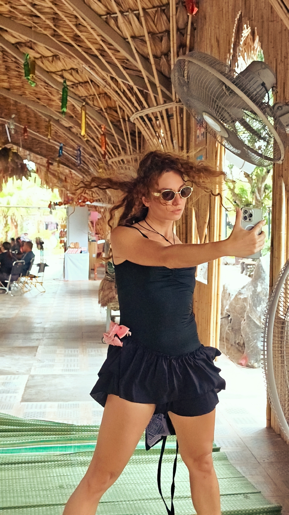 A woman with curly auburn hair, wearing sunglasses and a black tank top with a ruffled black skirt, takes a selfie with a smartphone in a covered outdoor area. Her hair is windblown. A pink accessory is clipped to her skirt. The area has a thatched roof supported by bamboo poles and decorated with colorful hanging ornaments. A large industrial fan is mounted on a bamboo wall. In the background, people are seated at tables, and there are market stalls with goods displayed on tables. The floor is tiled and a green mat is partially visible. The scene appears to be in a market or food stall area in Hoa Lư Ward, Vietnam, characterized by its tropical construction and vibrant decorations.