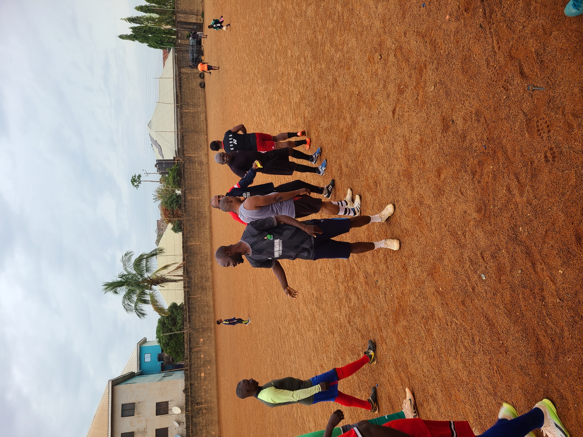 The image depicts a group of men on a reddish-brown dirt field in Barakin Lerawi, Nigeria. Several men are lined up and appear to be stretching or performing exercises. Another individual is running further away on the field. In the background, there are buildings, trees (including a palm tree), and a fence. The sky is overcast.