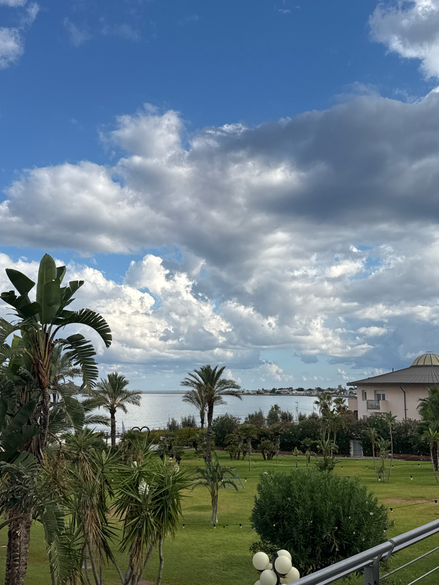 The image depicts a coastal landscape with a lush green lawn, palm trees, and a body of water in the background. The sky is partly cloudy, with both bright white cumulus clouds and darker, more ominous storm clouds.
In the foreground, on the left, a large Bird of Paradise plant with broad green leaves dominates the view, with several other palm trees of varying heights scattered across the lawn. The grass is neatly manicured, and string lights are visible hanging between some of the trees, suggesting an outdoor gathering space.
A building with a distinctive domed roof is situated on the right side of the frame, appearing to be a hotel or resort. The building has multiple stories and balconies. Beyond the lawn and trees, a calm body of water, likely the sea, stretches towards the horizon. A town or city skyline is faintly visible on the opposite shore. The overall impression is of a tropical or Mediterranean resort setting. The location is confirmed as being in the city of Capaci, Italy, a coastal town in Sicily known for its beaches and proximity to Palermo.