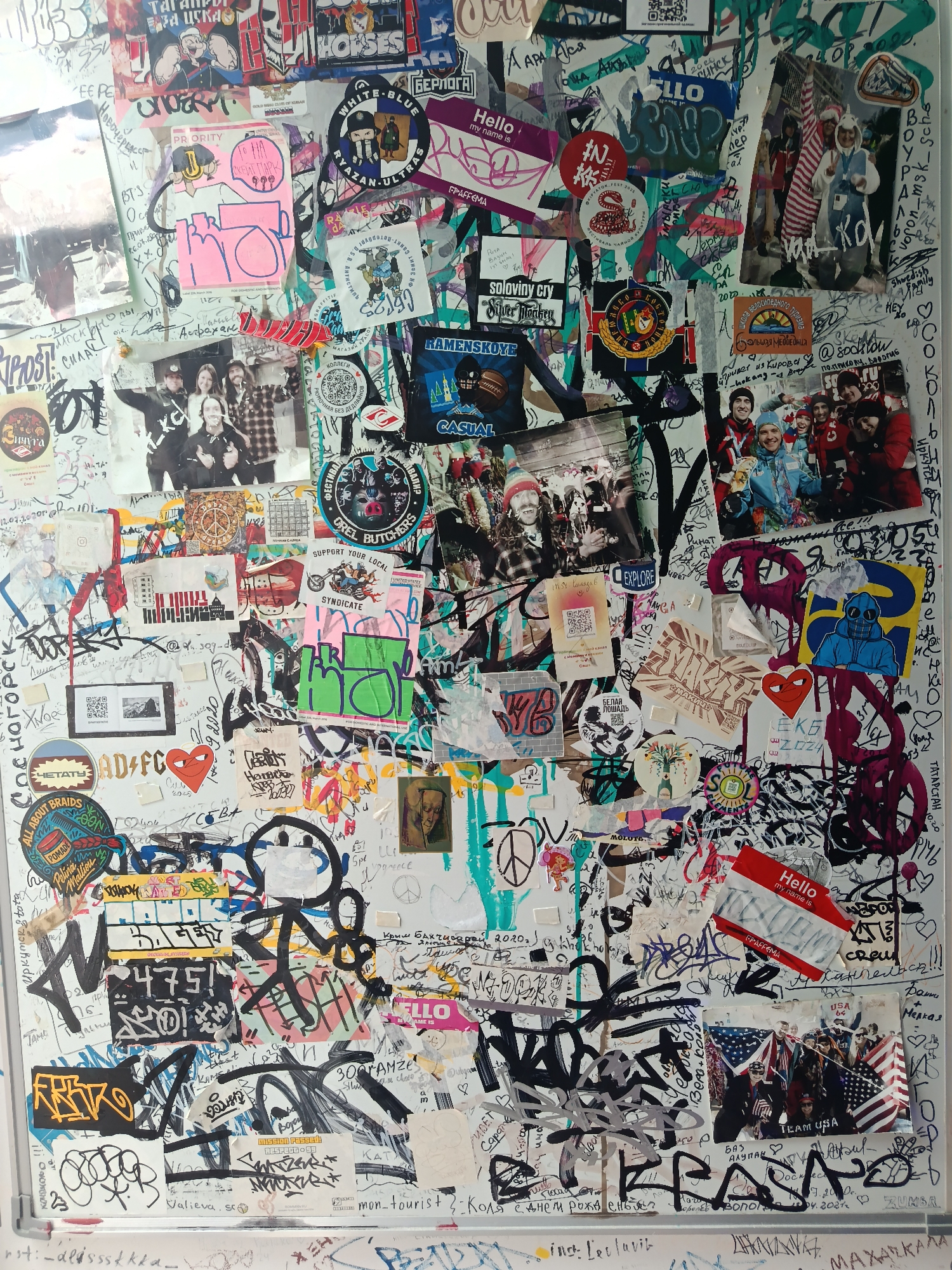 This media file is a photograph of a wall covered in a dense collage of stickers, posters, and graffiti. The subjects are varied, including numerous images of people, both in group photos and as individual portraits or stylized drawings. There are also various objects depicted, such as logos, symbols (like peace signs and hearts), and abstract graphic designs. The scene is chaotic and layered, suggesting a public space that has been heavily utilized for self-expression through street art and personal messages.
The overall impression is that of an informal bulletin board or a section of a skate park or youth hangout area where people have left their mark. There is no clear indication of a specific time of day or weather, but the diverse range of stickers, some with dates in the past (e.g., 2010, 2020, 2021), suggests this collection has accumulated over time. The location context provided is "Estosadok, Russia."
Notable details include a "Hello, my name is" sticker, numerous Cyrillic inscriptions and graffiti tags, and several stickers with specific locations or group names like "RYAZAN-ULTRAS," "OREL BUTCHERS," and "TEAM USA." There are also references to events and places like "SPYATOR FEST 2025" and "MOSCOW." The emotions conveyed are varied, from the playful and energetic in the cartoonish graphics to the more serious or defiant in the graffiti. Some images show people smiling and posing, hinting at positive social interactions. The wall itself is the primary setting, acting as a canvas for this vibrant display.
