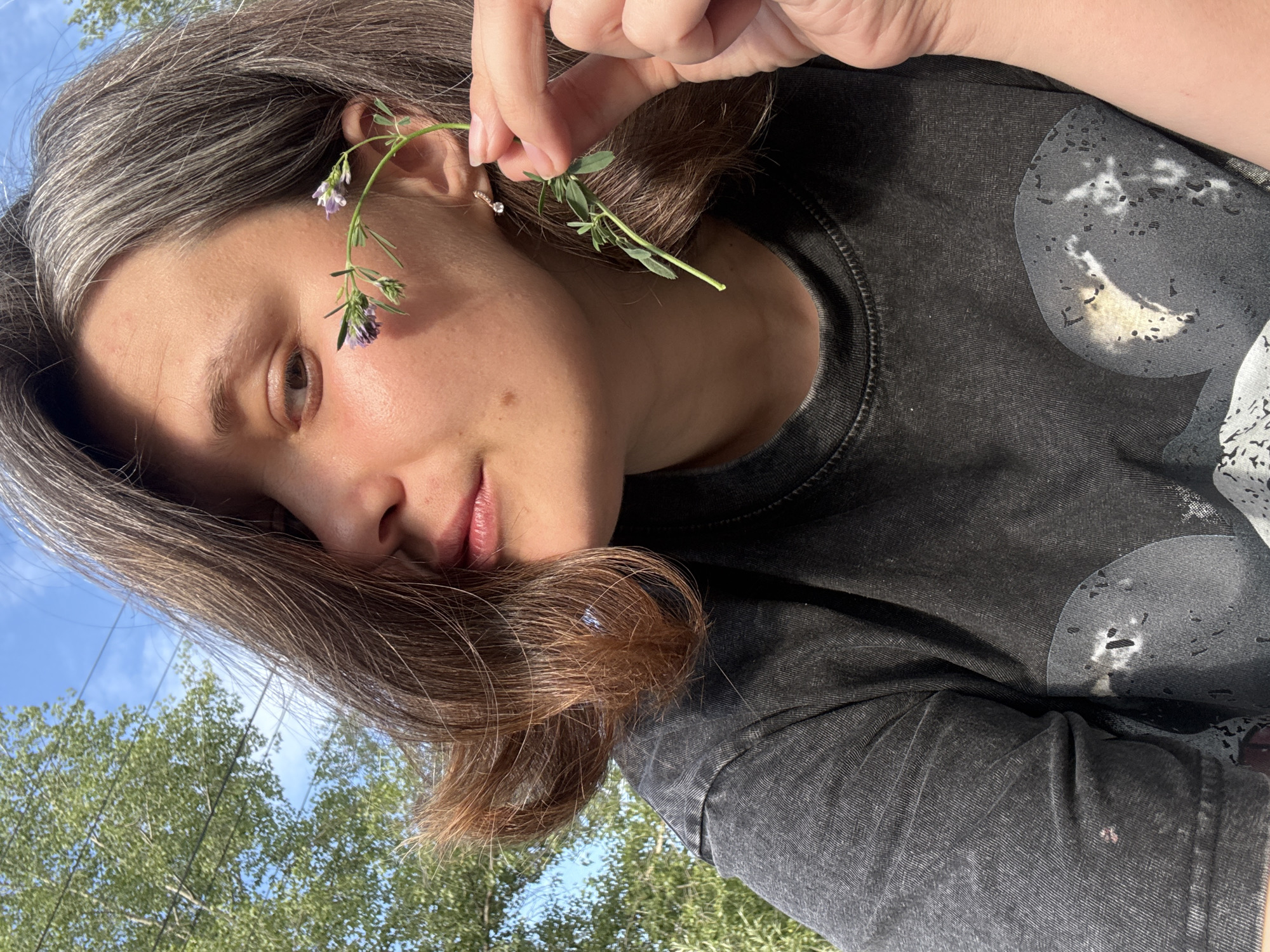 A young adult with brown hair, streaked with gray, is shown in a close-up, outdoor portrait. They are wearing a dark gray t-shirt. The subject is holding a delicate stem with small purple flowers and green leaves near their face and ear. Sunlight casts a shadow across their face, highlighting their cheek and eye. In the background, a large green tree with lush foliage is visible against a bright blue sky. The overall scene suggests a peaceful moment in nature, possibly in a park or garden. There is no specific indication of the city of Chaikovsky, Russia within the visual information provided.