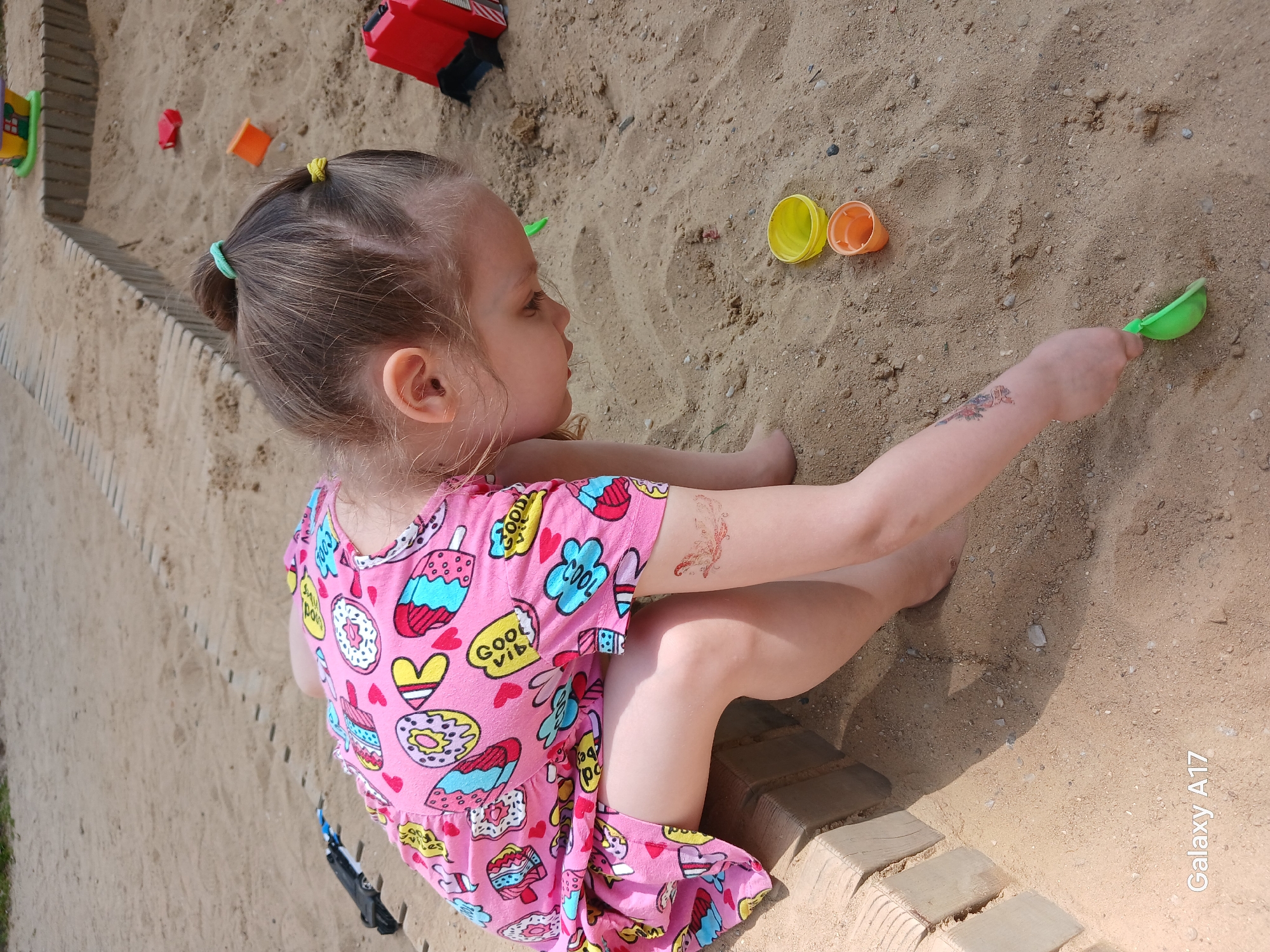A young girl is playing in a sand pit. The child has light brown hair in pigtails with a yellow and green hair tie, wearing a pink patterned dress. She is seated at the edge of the pit on a ledge. She is holding a green scoop in her hand. Orange and yellow plastic cups are in the sand next to her. Toys in red, green, and orange are visible in the sand as well. The location could be Yekaterinburg, Russia, or anywhere with a similar playground; however, there are no unique details to confirm this. The photo was taken on a Galaxy A17.